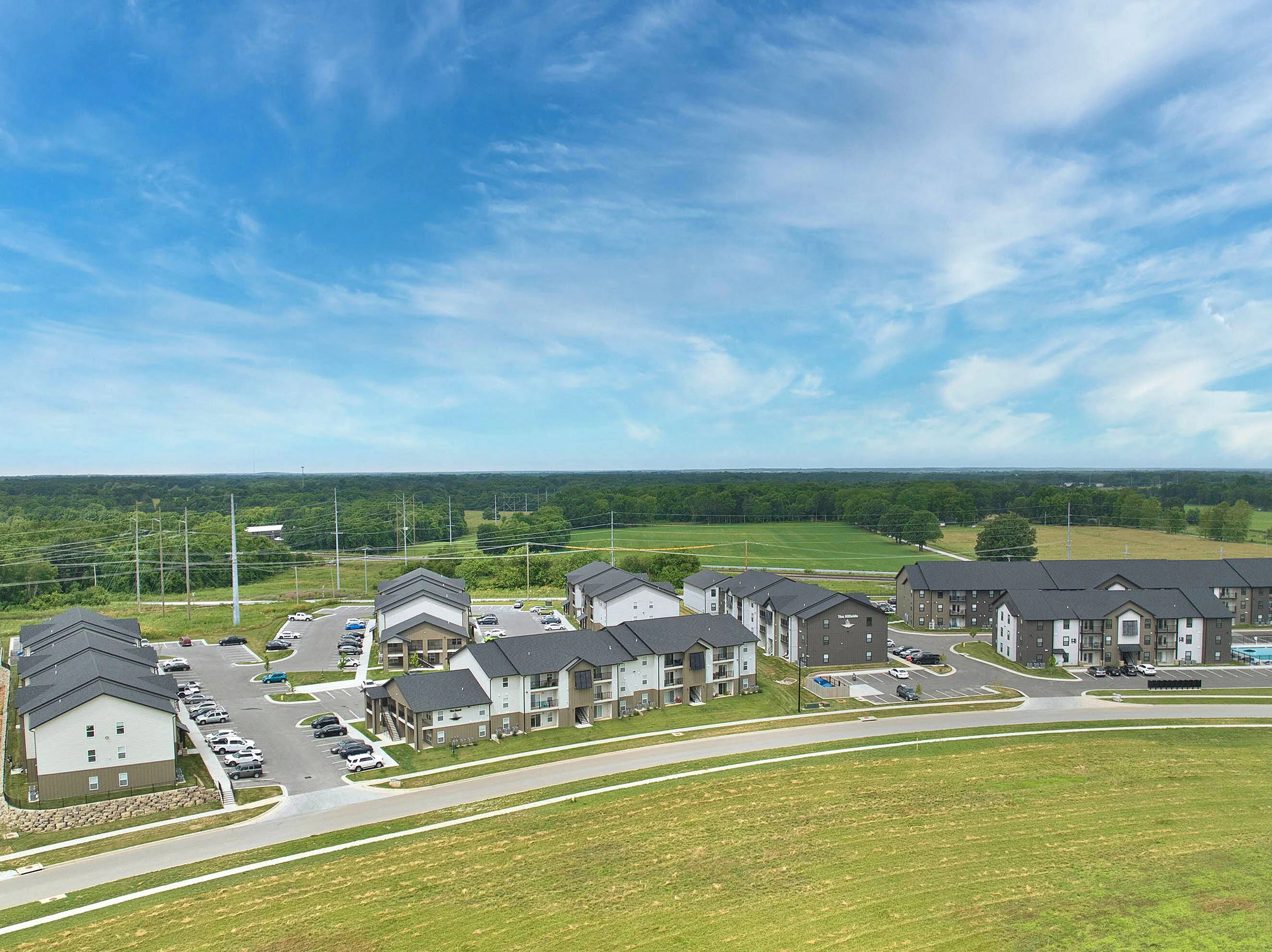 Scenic aerial view of modern apartment complex surrounded by lush greenery and open fields under a vibrant blue sky.
