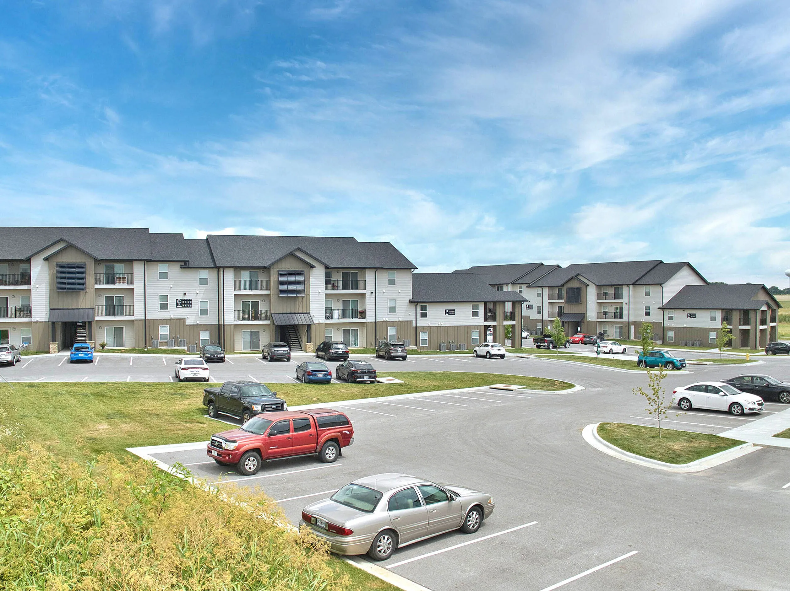Modern apartment complex featuring spacious buildings with balconies, ample parking, and scenic views of a bright blue sky.