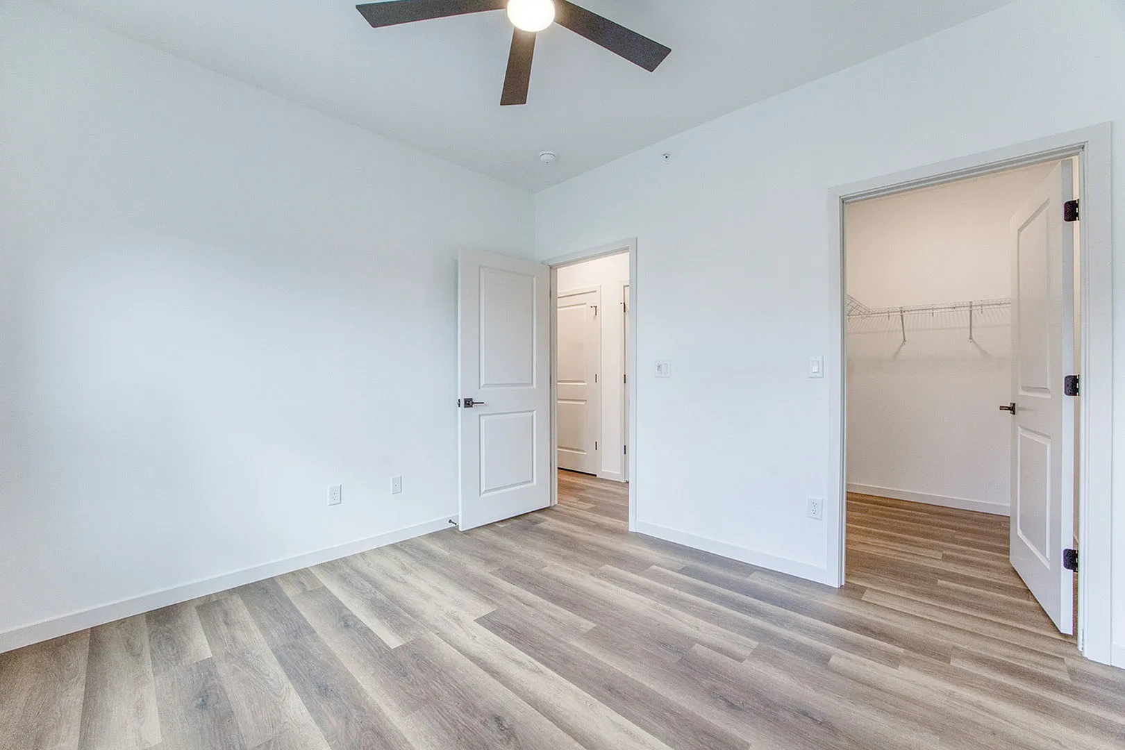 Bright bedroom with elegant wood flooring and ceiling fan, featuring two open doors leading to a spacious closet.