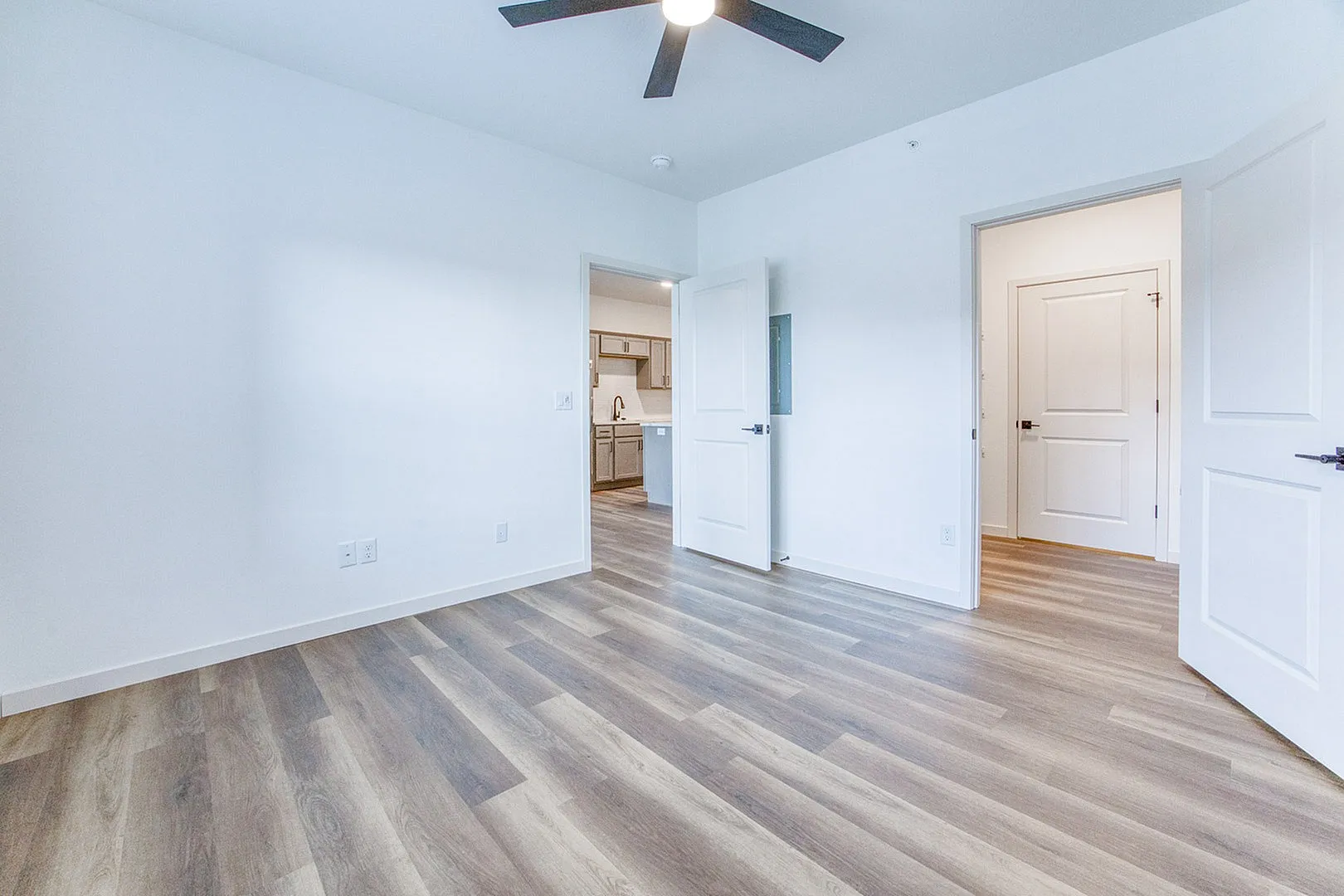 Sleek modern apartment room with elegant wood flooring, bright natural lighting, and a ceiling fan, offering a view into a contemporary kitchen.