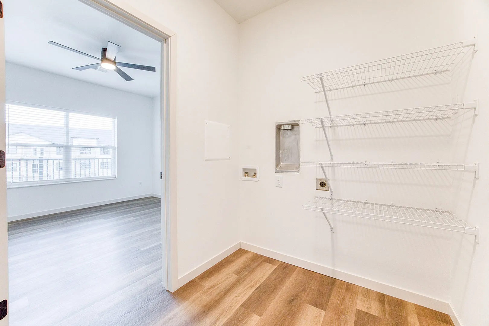 Bright, modern room with hardwood floors, large window, ceiling fan, and wire shelving, ideal for versatile living.