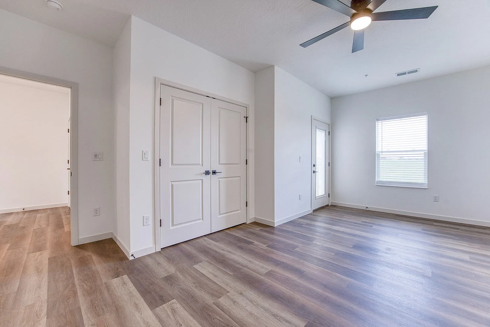 Bright and spacious room with elegant wood flooring, modern ceiling fan, and ample natural light from large window and glass door.