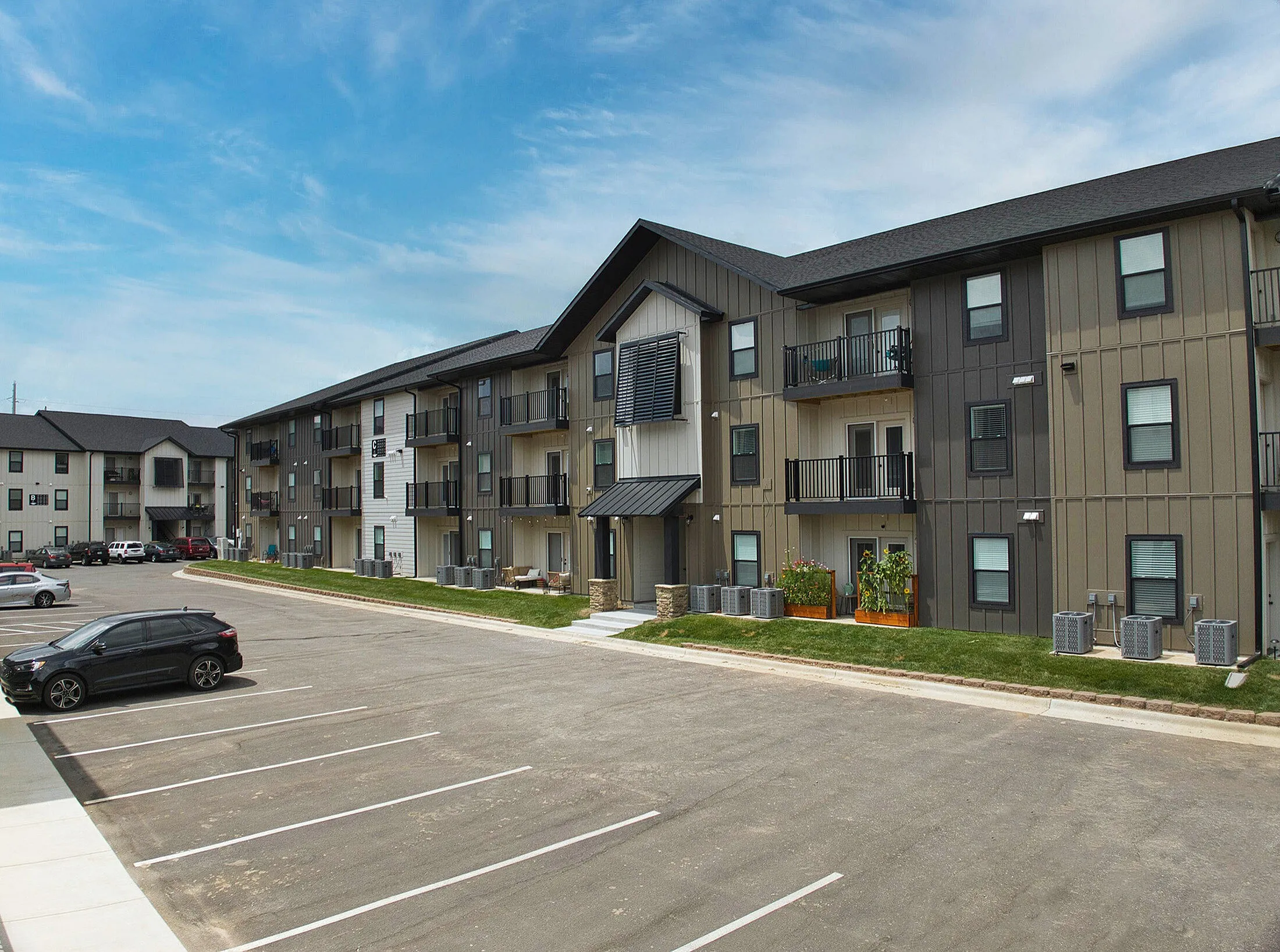 Modern three-story apartment building with balconies, ample parking space, and manicured green surroundings under a clear blue sky.