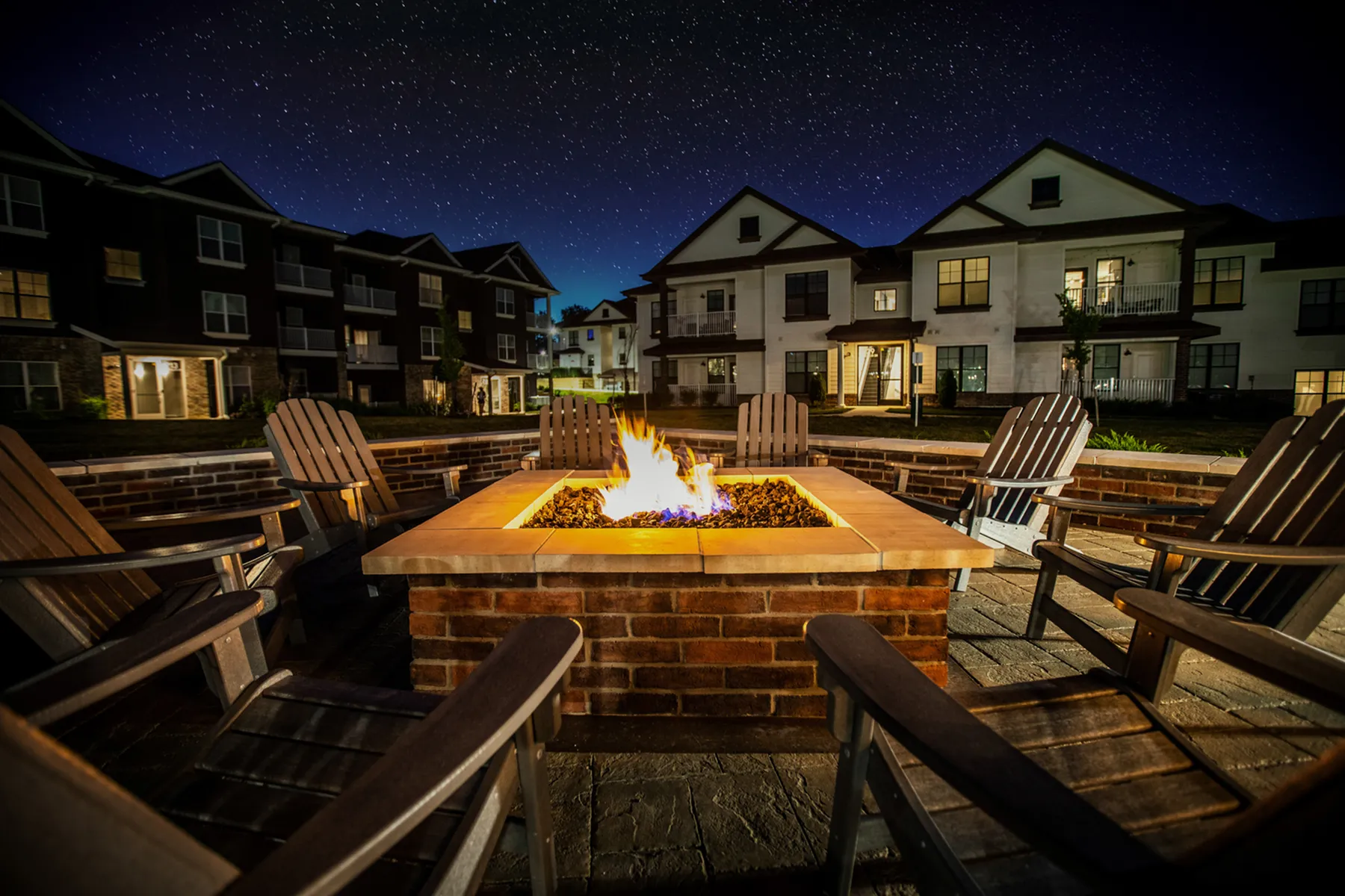 Captivating nighttime view of luxury apartment complex featuring an inviting fire pit surrounded by cozy chairs under a starry sky.