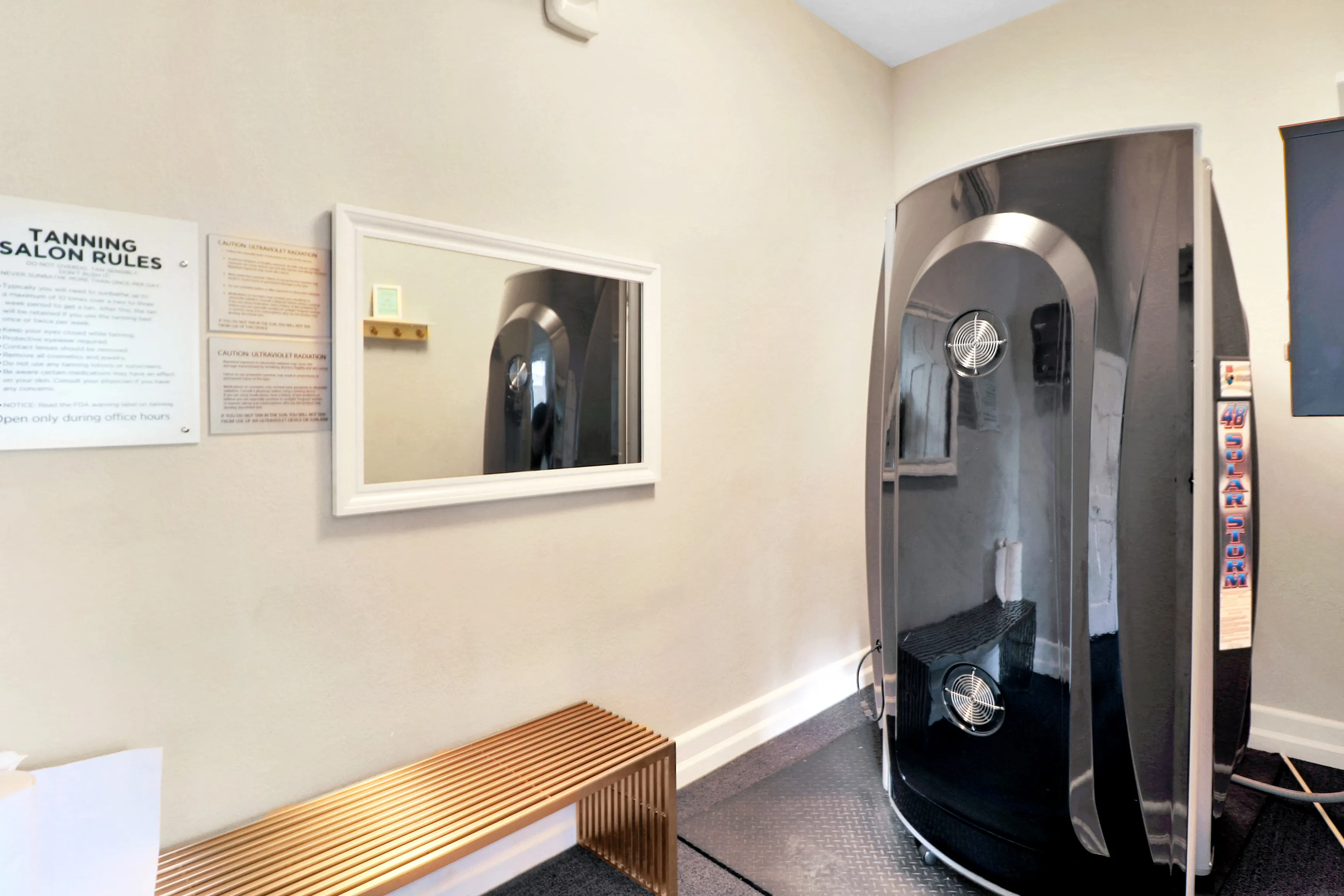 Modern tanning salon room featuring sleek tanning booth and stylish bench with a large mirror and informational signage on the wall.