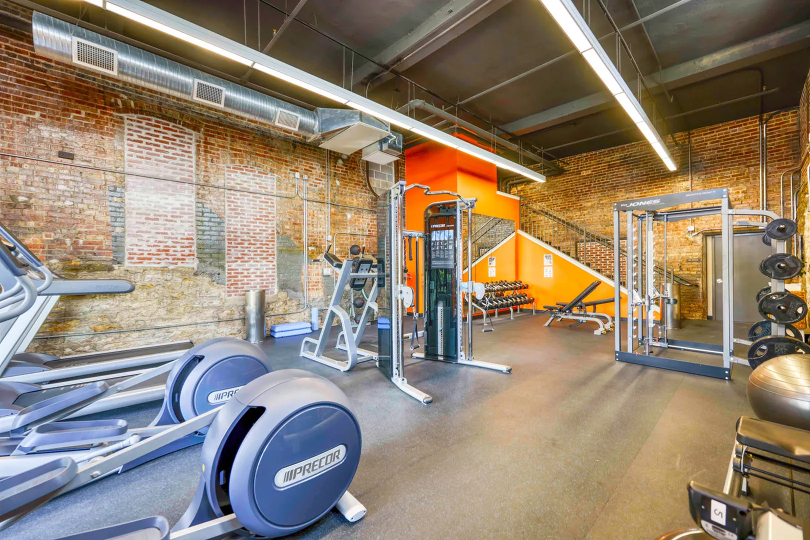 Modern gym with exposed brick walls, complete with state-of-the-art exercise machines, weights, and vibrant orange accents for an energetic workout environment.