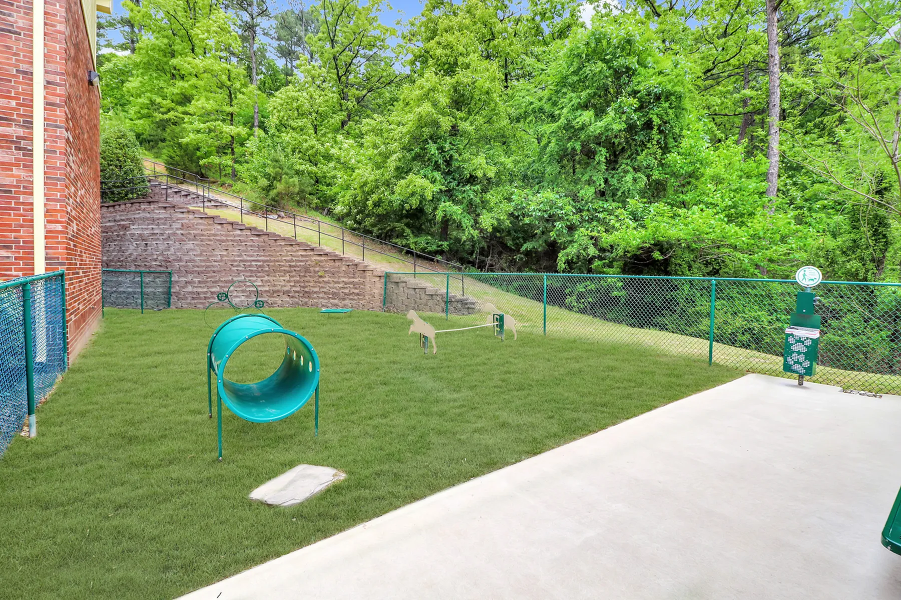 Charming apartment backyard featuring a fenced pet play area with a vibrant green lawn, agility equipment, and lush forest views.