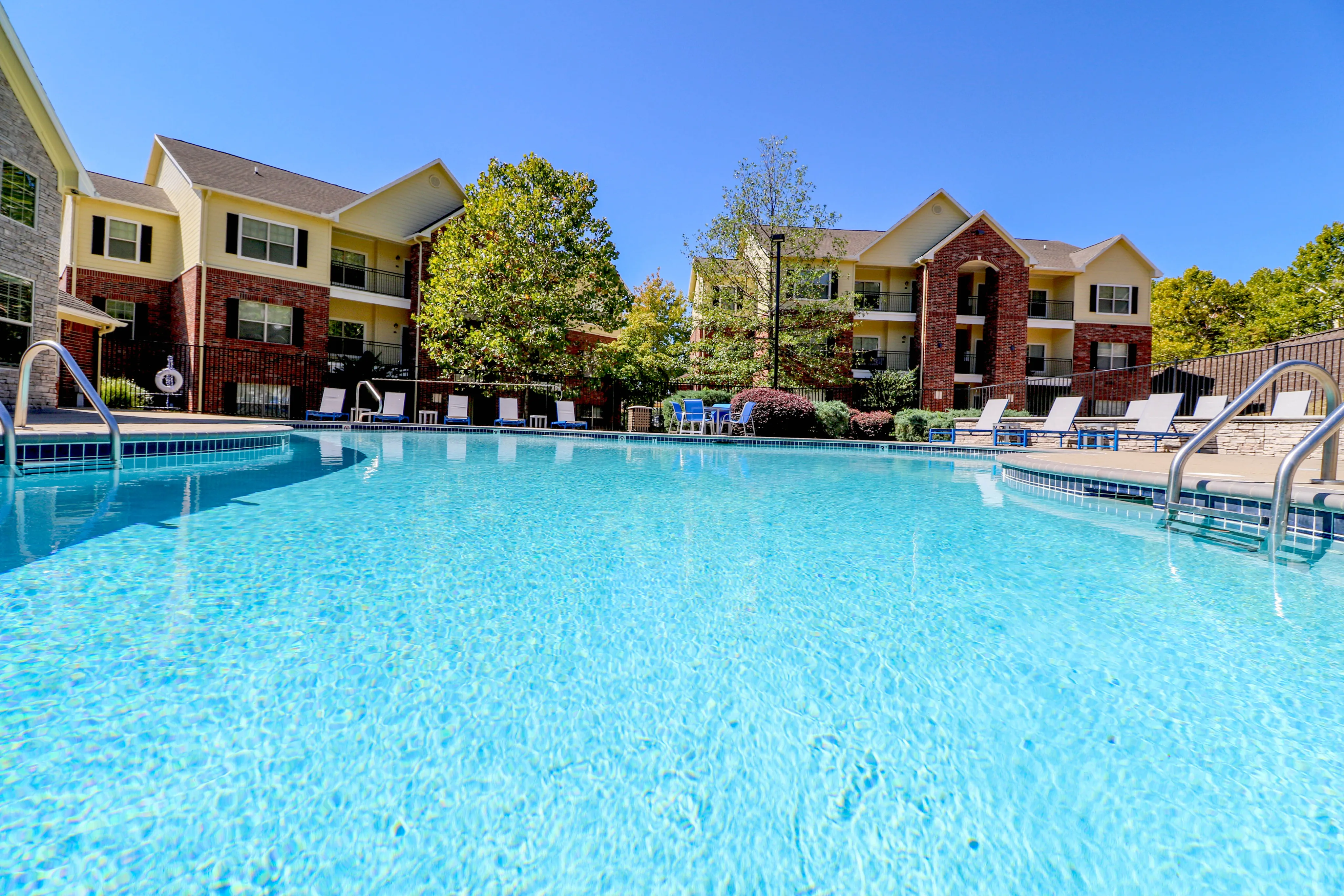 Inviting apartment complex featuring a sparkling outdoor pool, lounge chairs, and lush greenery under a clear blue sky.