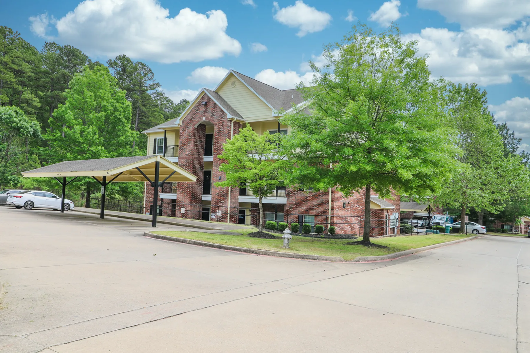 Charming red-brick apartment complex surrounded by lush greenery, featuring convenient covered parking and a welcoming community setting under a vibrant blue sky.