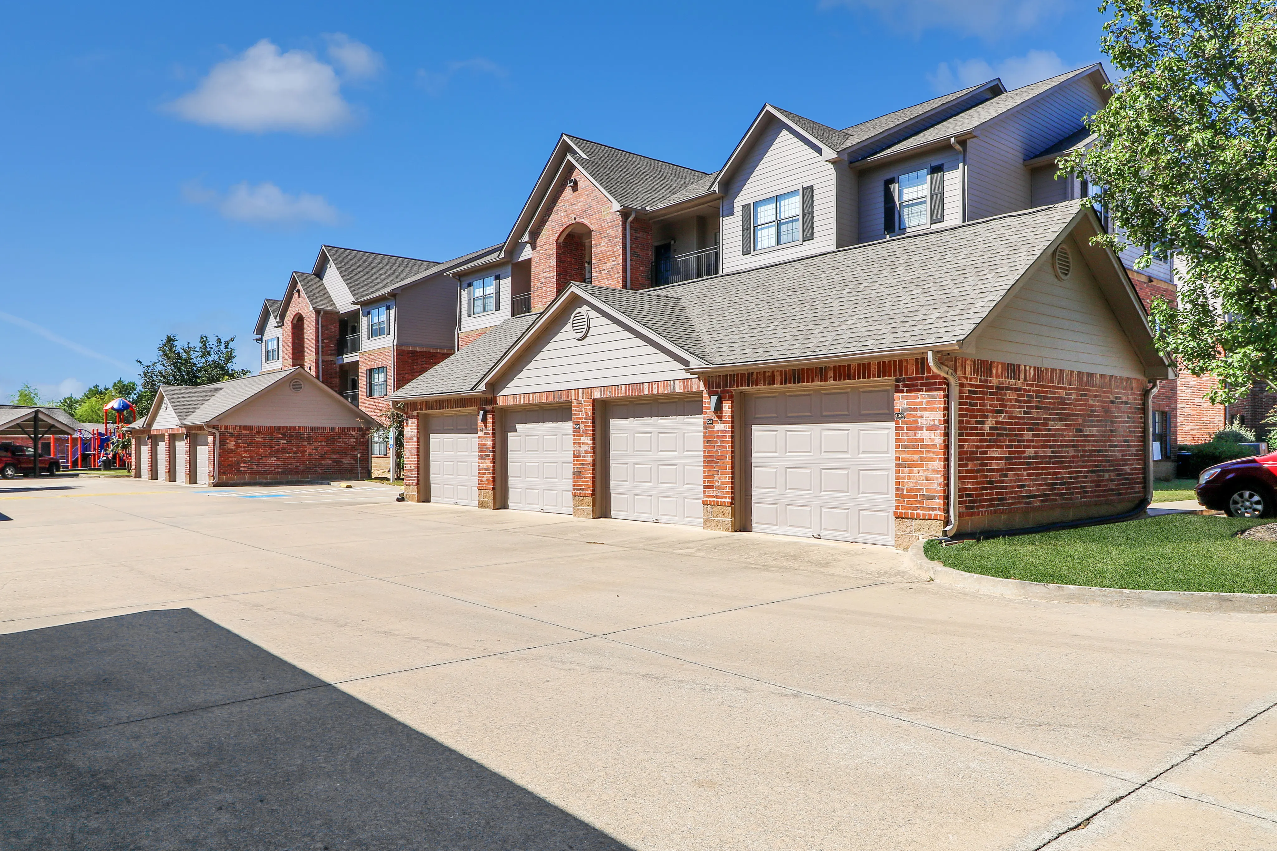 Inviting apartment community with attached garages, charming brick facade, and playground under a sunny sky.