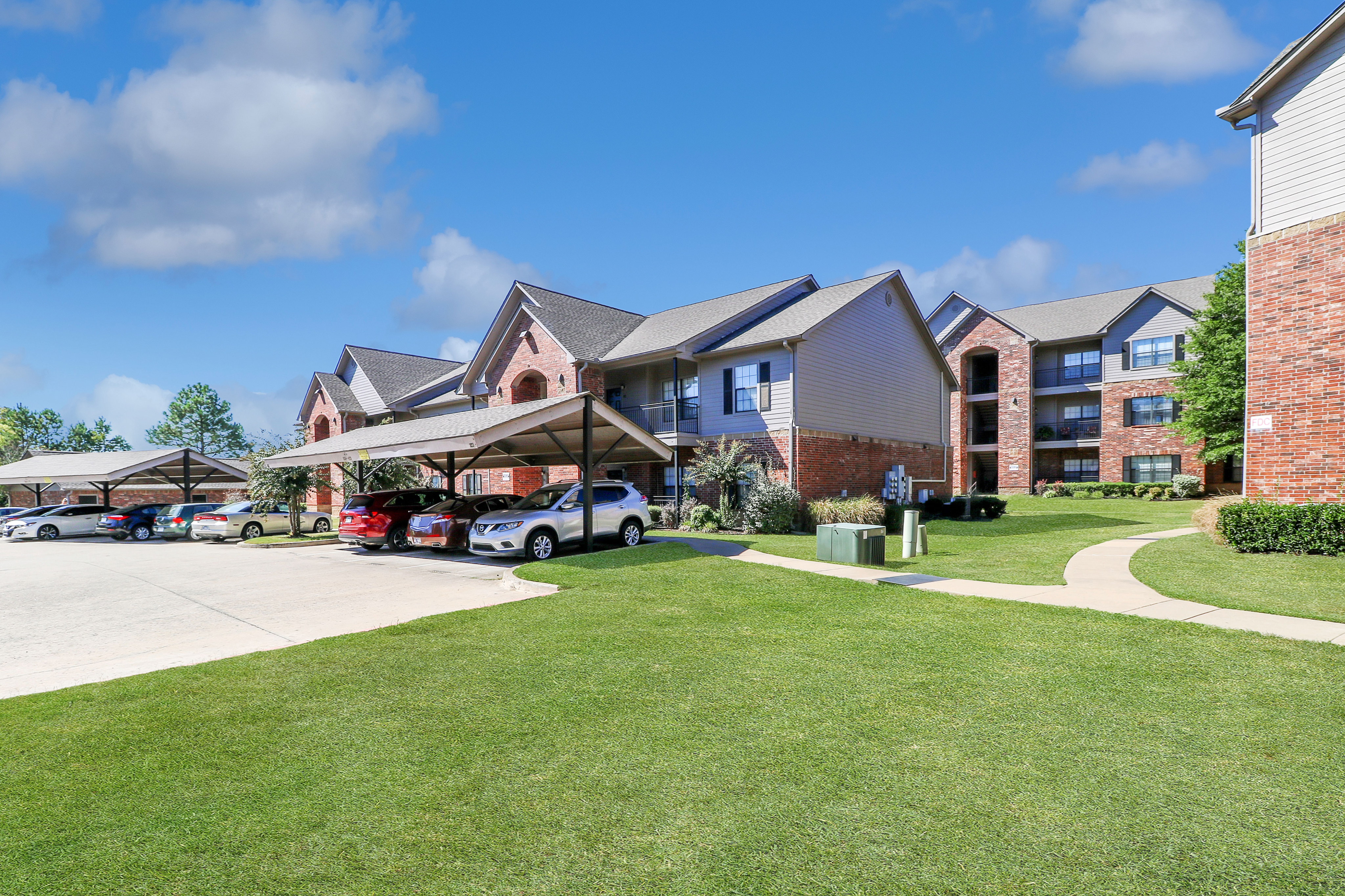 Inviting suburban apartment complex with neat brick exteriors, lush green lawns, and convenient carport parking under a bright blue sky.
