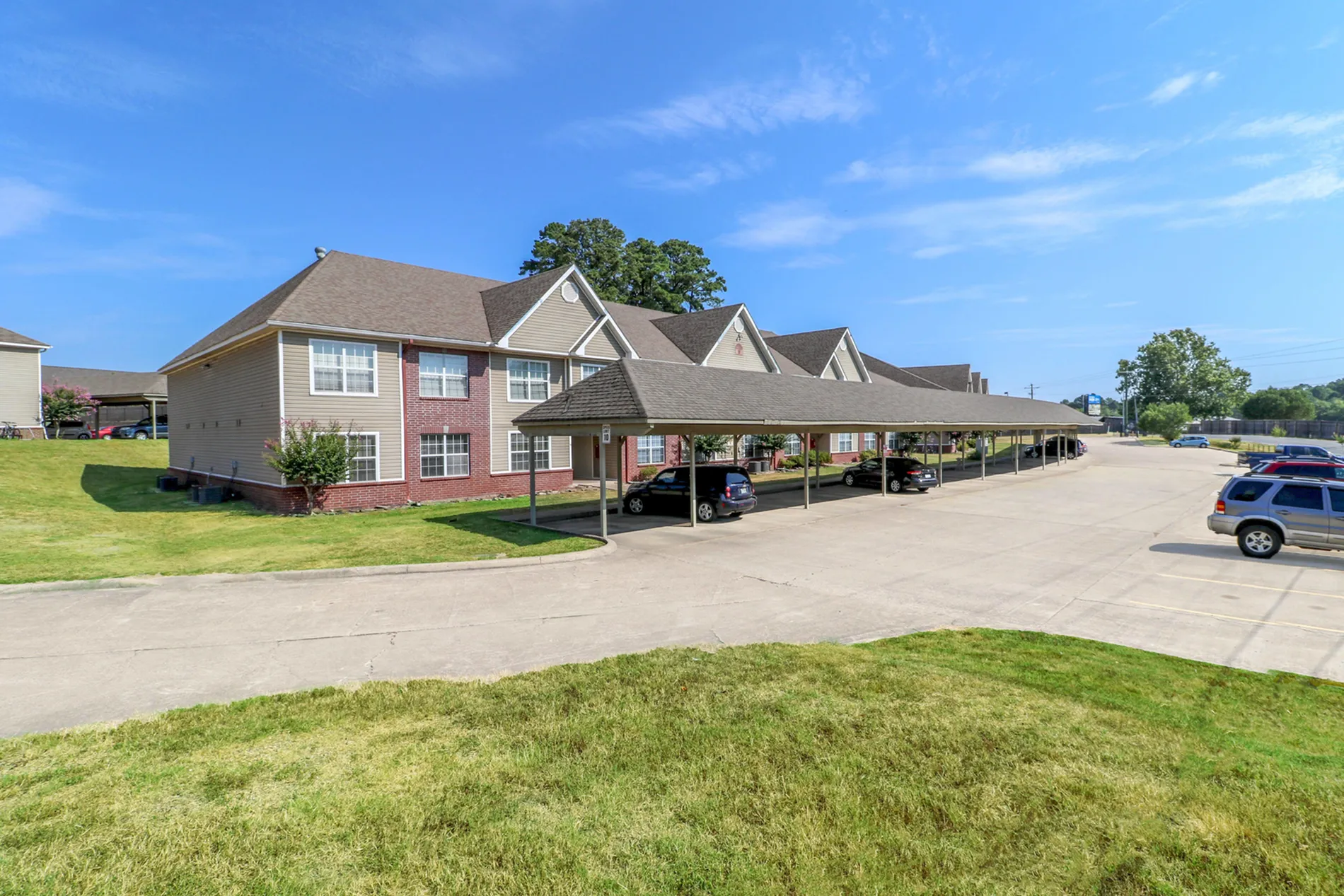 Charming apartment complex exterior featuring dual-story design with brick accents, covered parking, and beautifully landscaped greenery under a clear blue sky.
