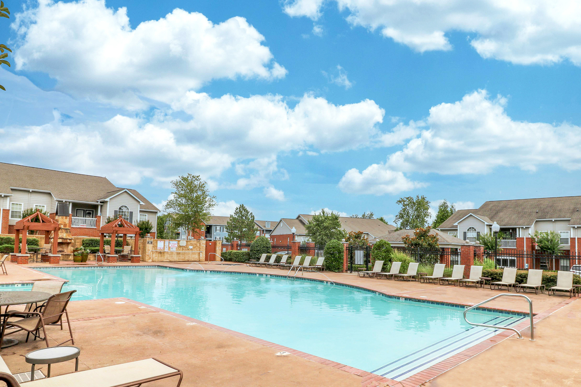 Inviting apartment complex with a large, sparkling swimming pool surrounded by lounge chairs, lush landscaping, and charming brick architecture under a bright blue sky.