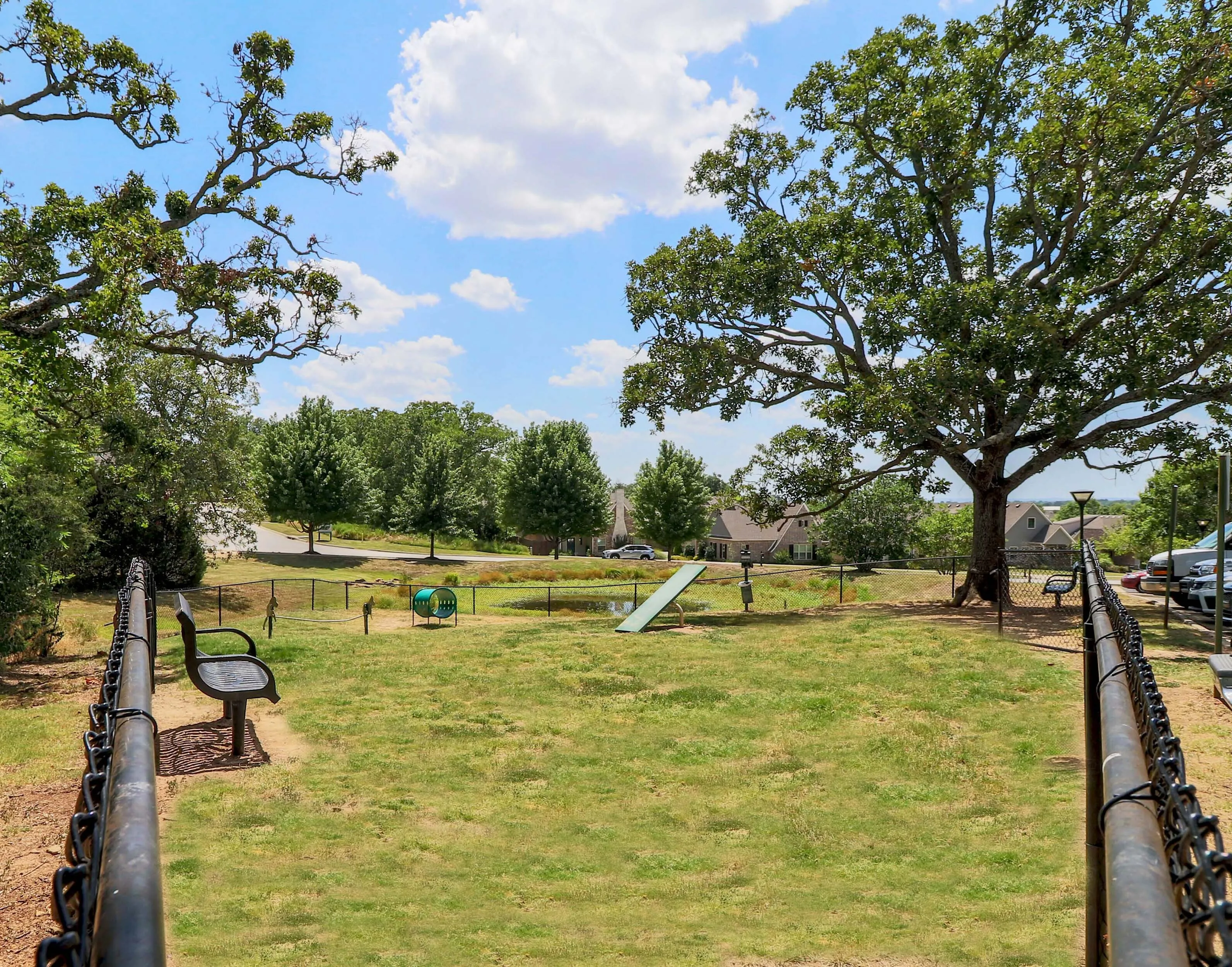 Charming fenced dog park with lush greenery, benches, and a picturesque neighborhood backdrop under a sunny blue sky.
