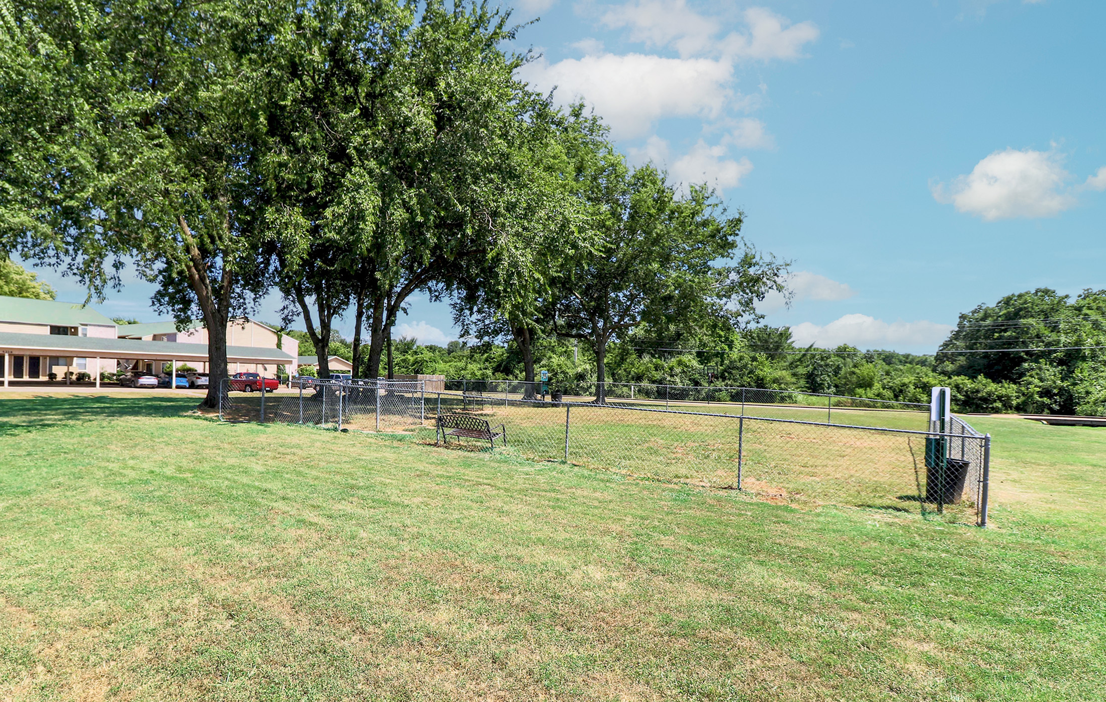 Expansive green recreational area with mature trees providing shade, enclosed by a fence for privacy and safety, ideal for outdoor relaxation and activities.