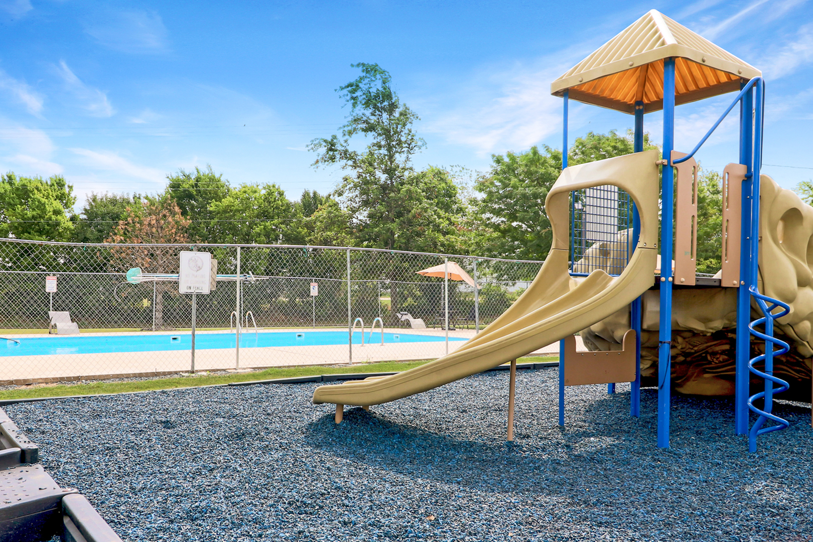 Inviting outdoor playground with a slide beside a sparkling swimming pool, surrounded by lush green trees and clear blue skies.
