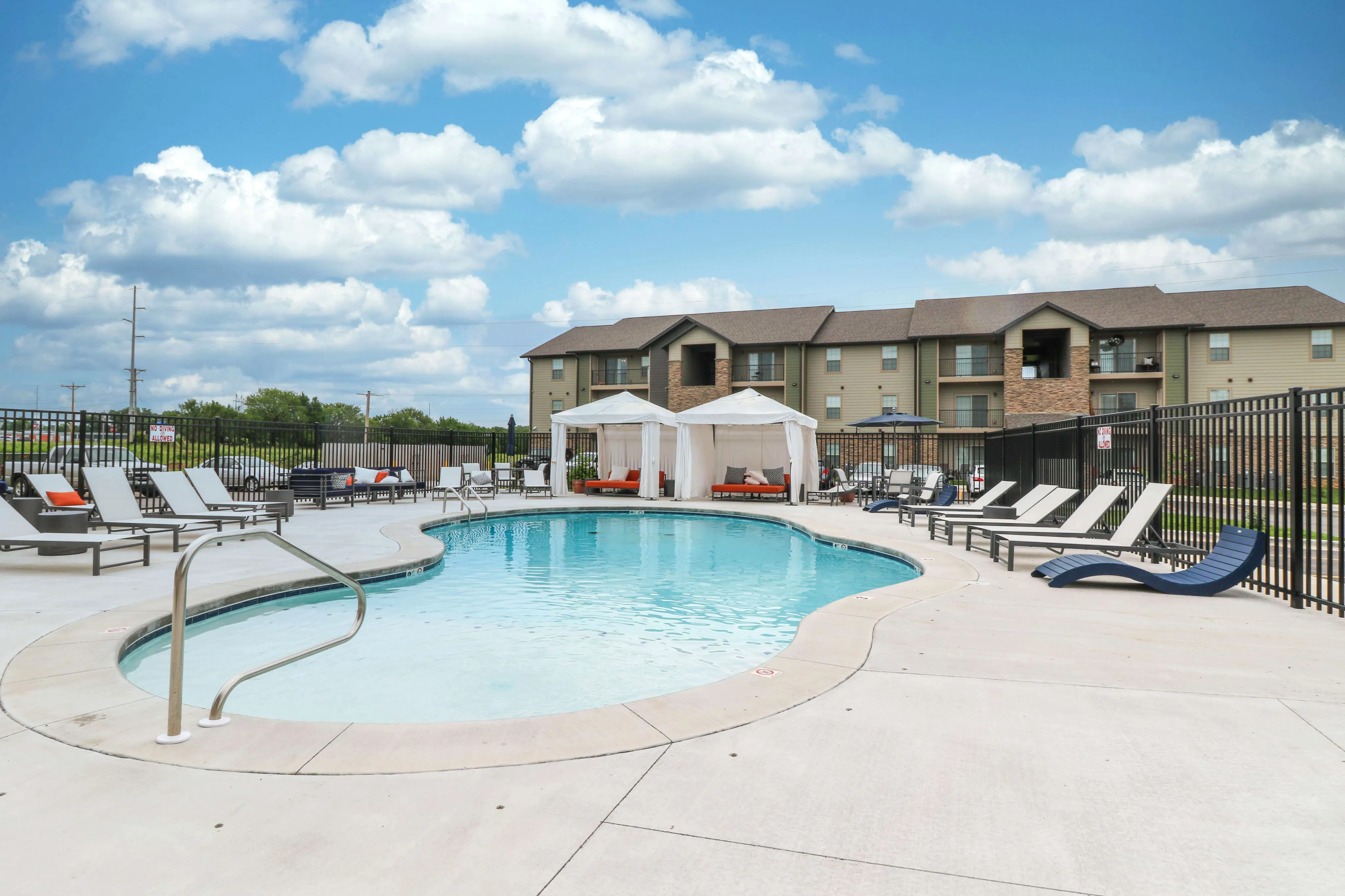 Inviting apartment complex with a luxurious swimming pool area surrounded by comfortable loungers and elegant cabanas under a clear blue sky.