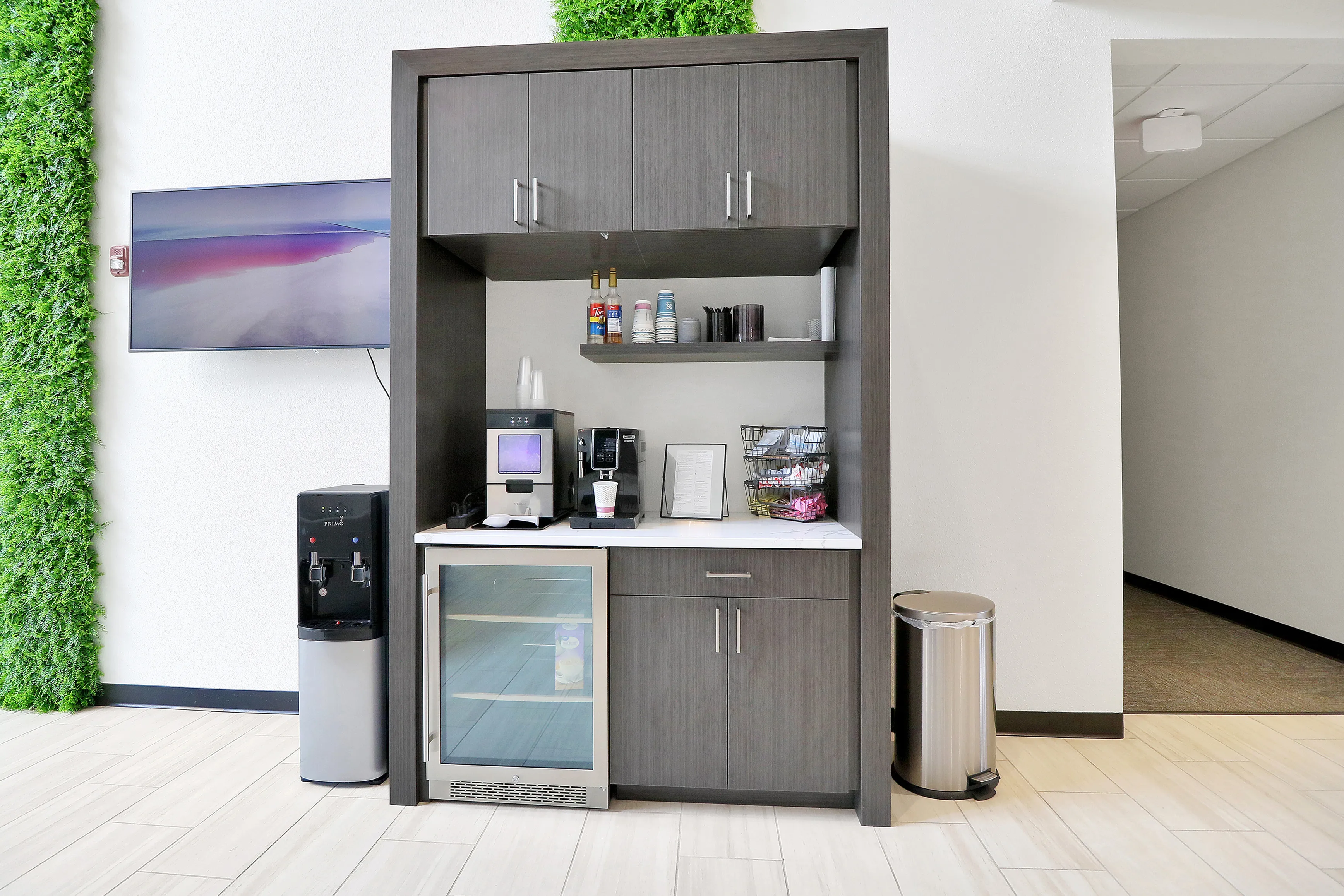 Modern refreshment nook with sleek cabinetry, featuring a coffee machine, beverage fridge, and a wall-mounted TV, ideal for relaxed living.