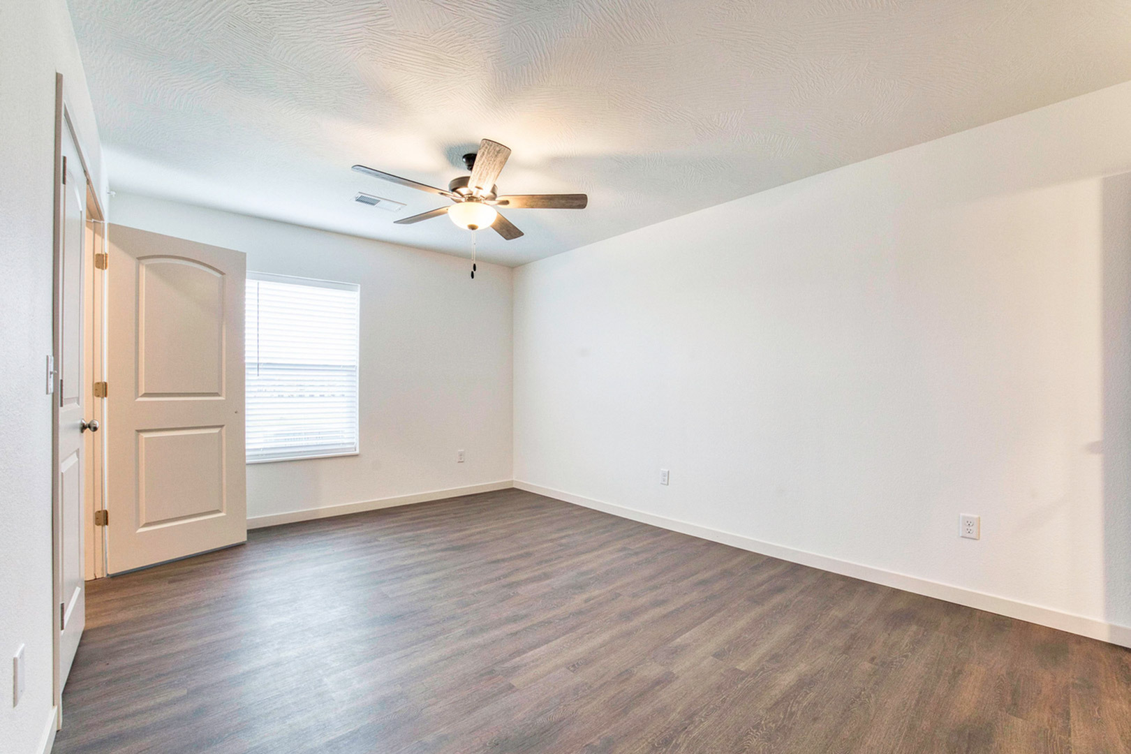 Spacious and airy empty room with elegant wood flooring, a ceiling fan, and ample natural light through large window.
