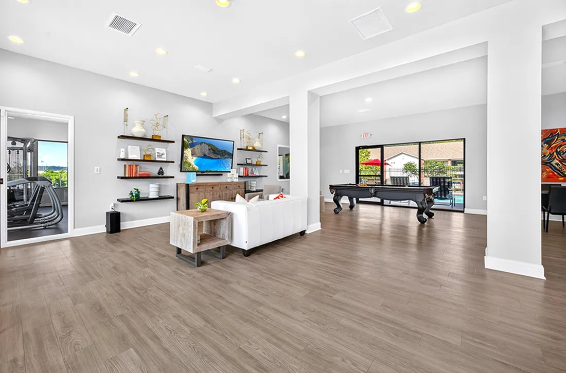 Contemporary living space with stylish wooden floors, sleek modern shelving, and cozy seating, featuring a large TV and pool table with natural light streaming through glass doors.