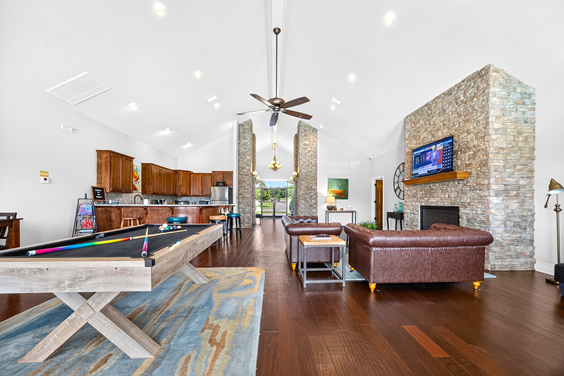 Elegant open-concept living room with stone fireplace, modern kitchen, and stylish pool table, perfect for entertainment.