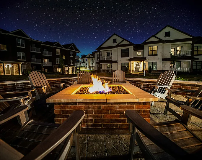 Nighttime view of a lit fire pit surrounded by Adirondack chairs, with apartment buildings in the background and a starry sky at Meridian at View High Apartments in Lee&rsquo;s Summit, Missouri.