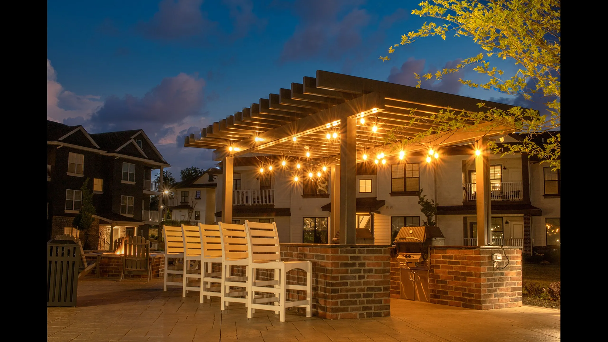 Grilling station with bar seating and overhead lighting, firepit in the background, and apartment buildings behind at dusk at Meridian at View High Apartments in Lee&rsquo;s Summit, Missouri.