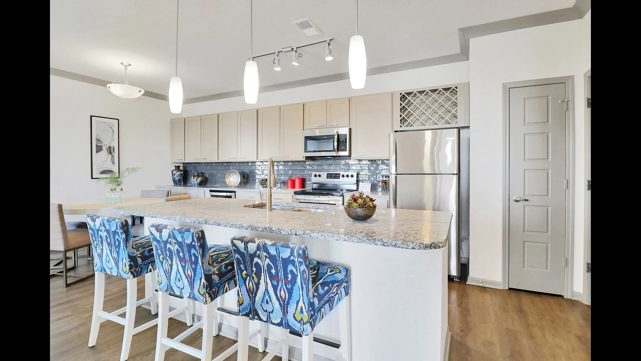 Modern kitchen with center island, hardwood floors, stainless steel appliances, and sleek cabinetry, with dining and living areas visible in the background at Meridian at View High Apartments in Lee&rsquo;s Summit, Missouri.