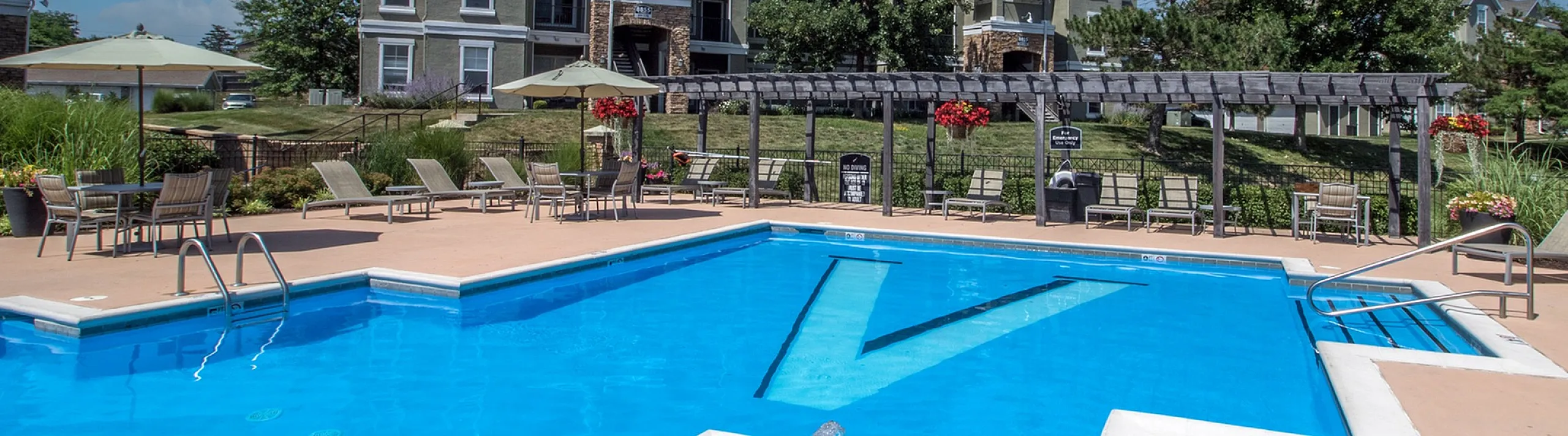 Luxurious apartment complex with a stunning outdoor pool, surrounded by lush greenery and comfortable lounging chairs under a bright blue sky.
