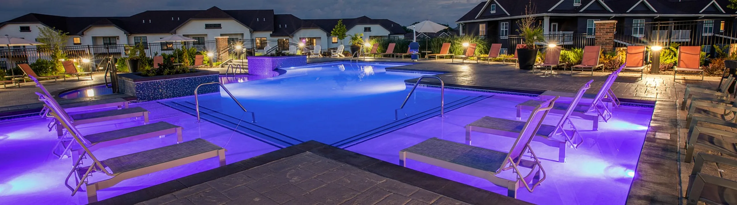 Elegant apartment complex featuring illuminated pool with modern lounge chairs and vibrant evening sky backdrop.