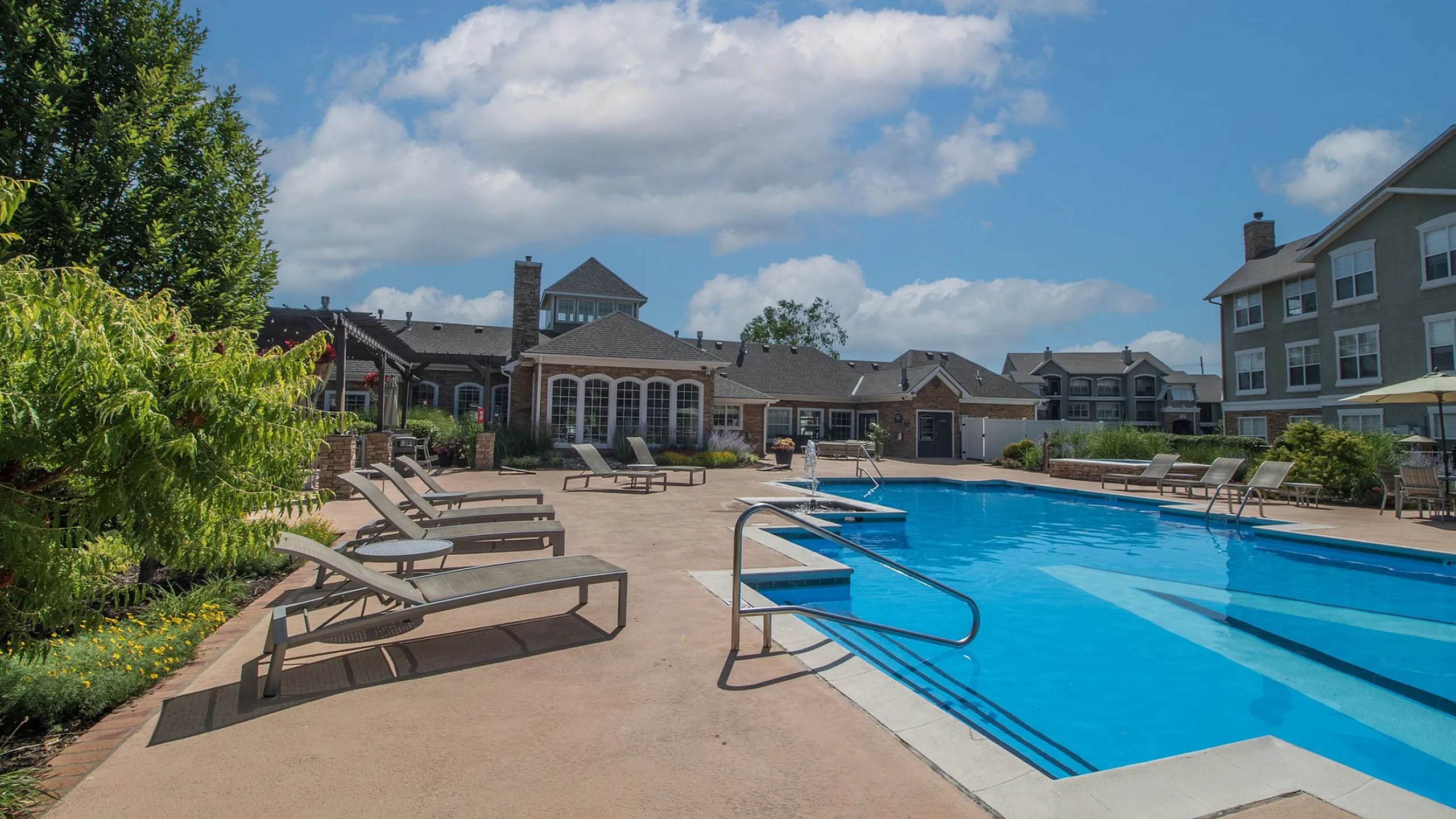 Inviting apartment complex pool area with elegant lounge chairs, surrounded by manicured greenery and charming architecture under a bright blue sky.