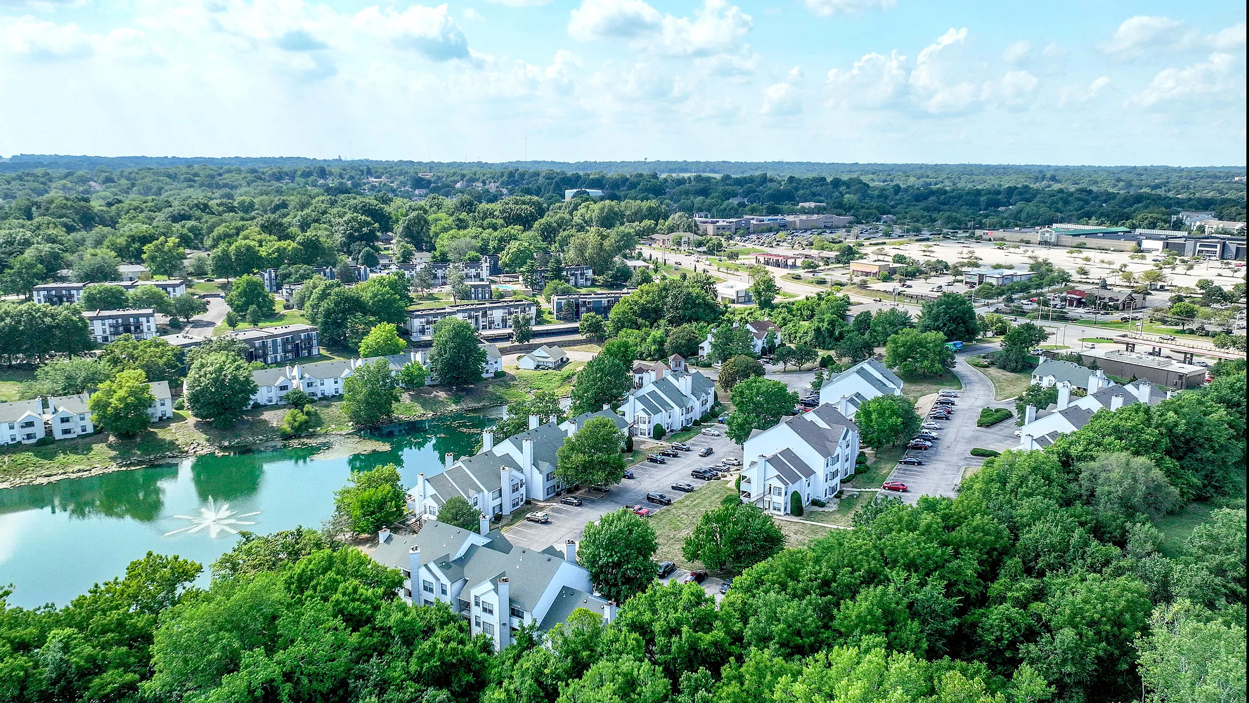 Aerial view of charming apartment complex surrounded by lush greenery and a serene pond, offering a peaceful living environment with convenient access to amenities.