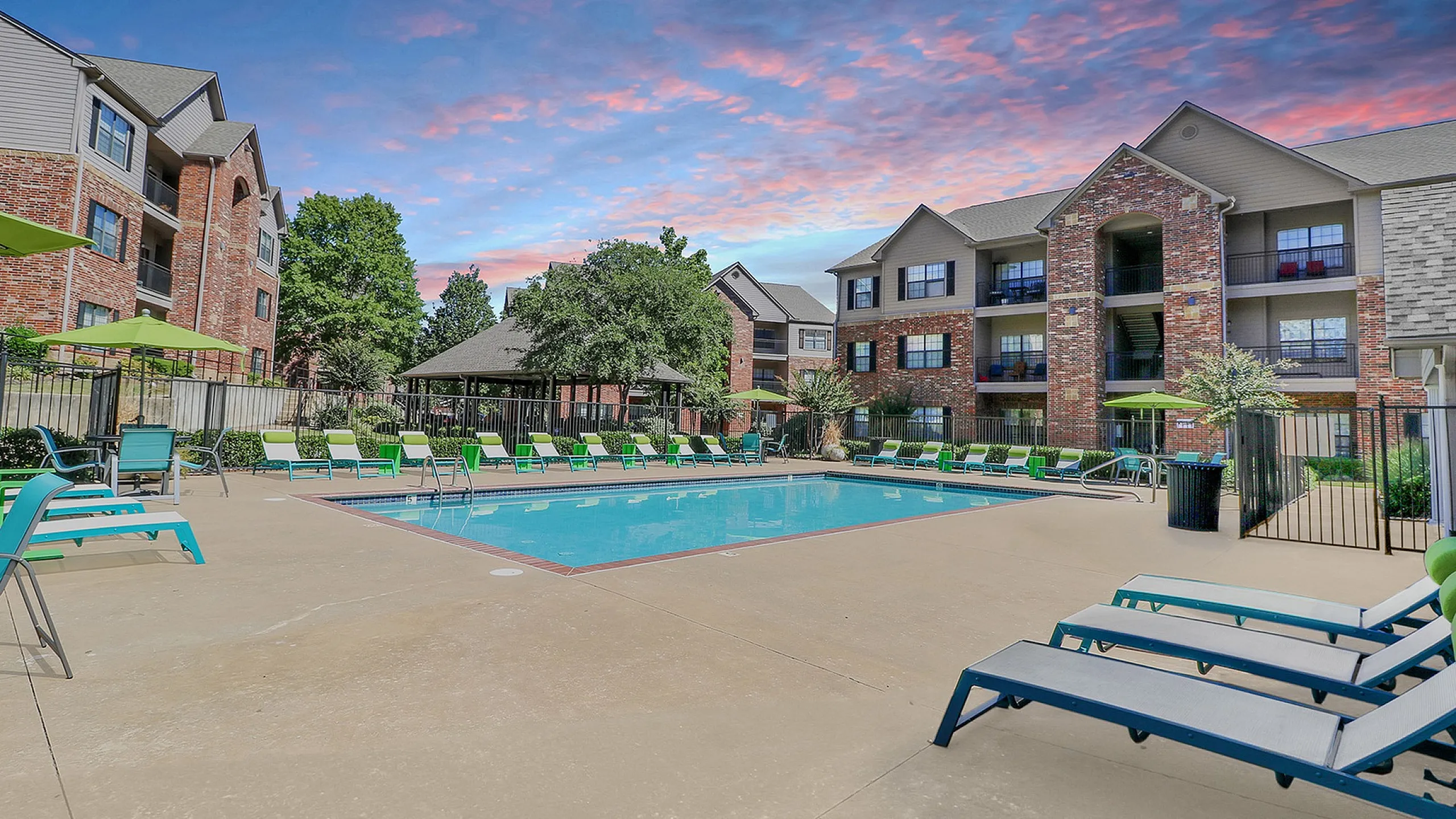 Elegant apartment complex with a sparkling outdoor pool, surrounded by lush greenery and stylish lounge chairs, set against a stunning sunset sky.