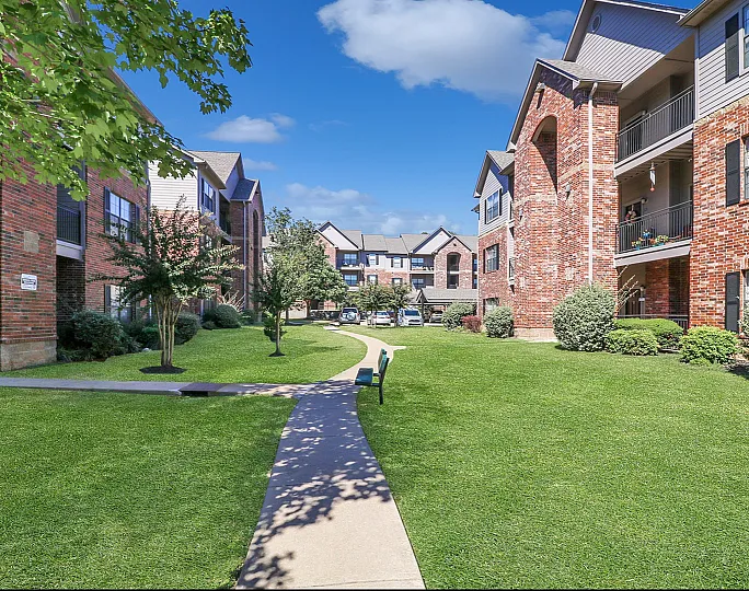 Inviting apartment courtyard with lush green lawns, brick buildings, and a peaceful walkway under a bright blue sky.