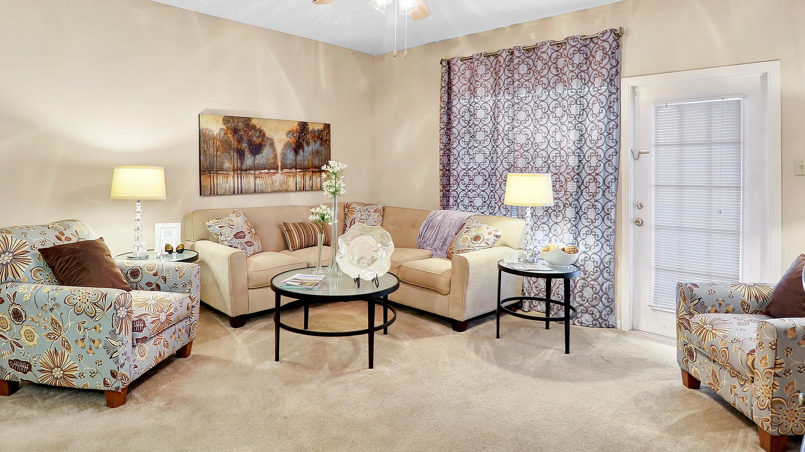 Inviting living room with plush beige sofas, floral armchairs, and a chic ceiling fan, bathed in warm lighting and complemented by elegant decor.