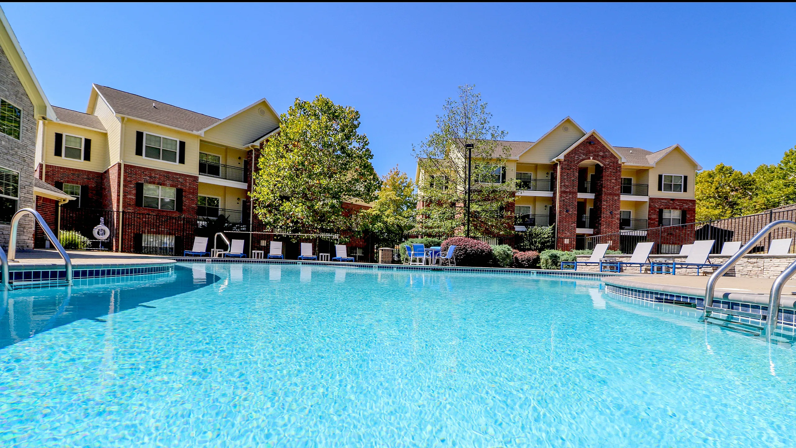 Inviting apartment complex with a sparkling outdoor swimming pool, surrounded by sun loungers and lush greenery.