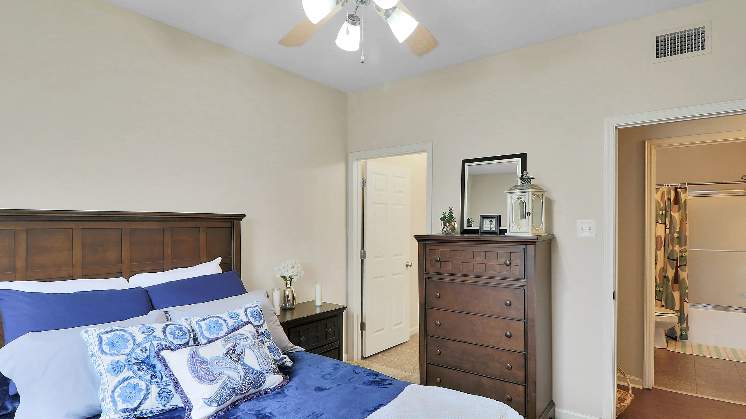 Inviting bedroom with elegant wooden furniture, plush bedding in soothing blue tones, soft lighting from a ceiling fan, and an en-suite bathroom for added convenience.
