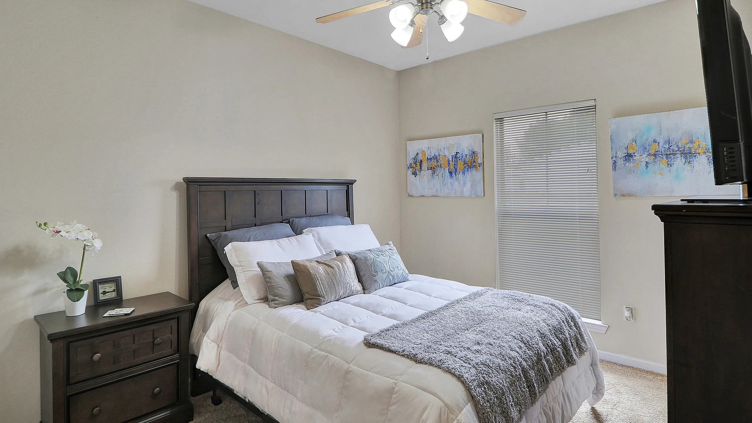 Inviting bedroom with a cozy double bed, warm lighting from an elegant ceiling fan, and tasteful wall art, offering a serene and stylish retreat.