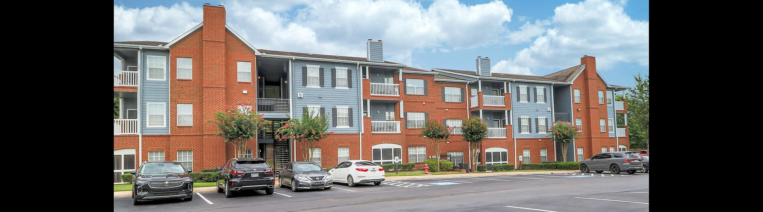 Charming brick apartment complex with three stories, featuring spacious balconies and ample parking under a bright blue sky.