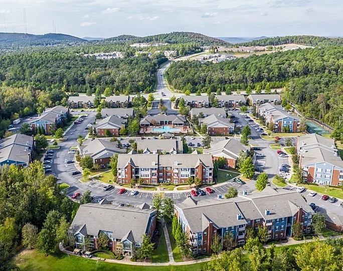 Aerial view of a charming apartment community nestled in lush green surroundings, featuring modern buildings with ample parking and scenic pathways.
