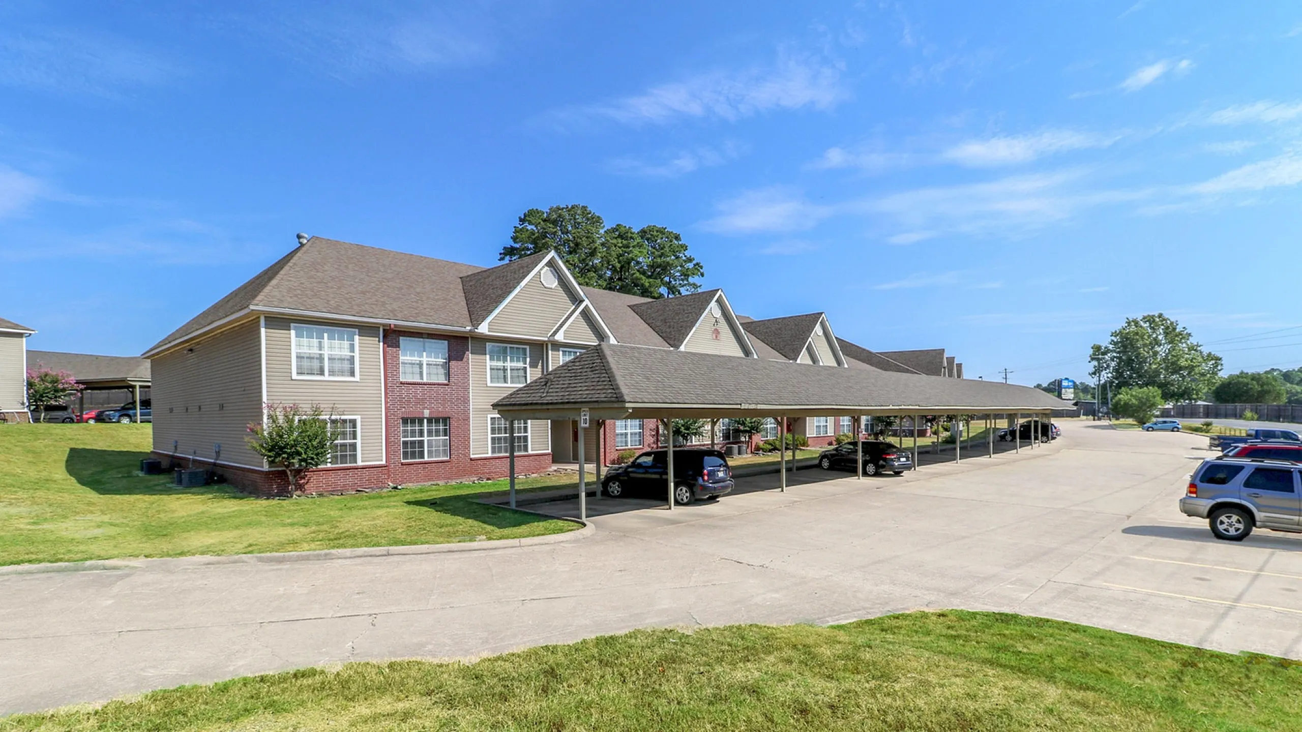Charming two-story apartment complex with brick and beige exterior, featuring covered parking and lush green surroundings under a bright blue sky.