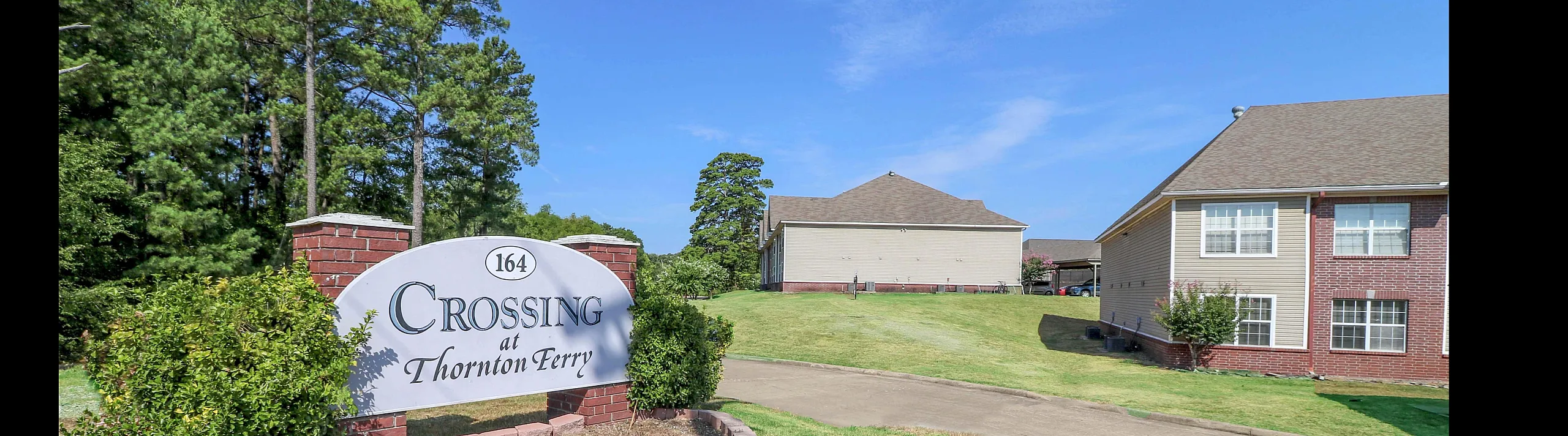 Welcoming entrance to the Crossing at Thornton Ferry surrounded by lush green lawns and scenic trees, showcasing a serene residential community.