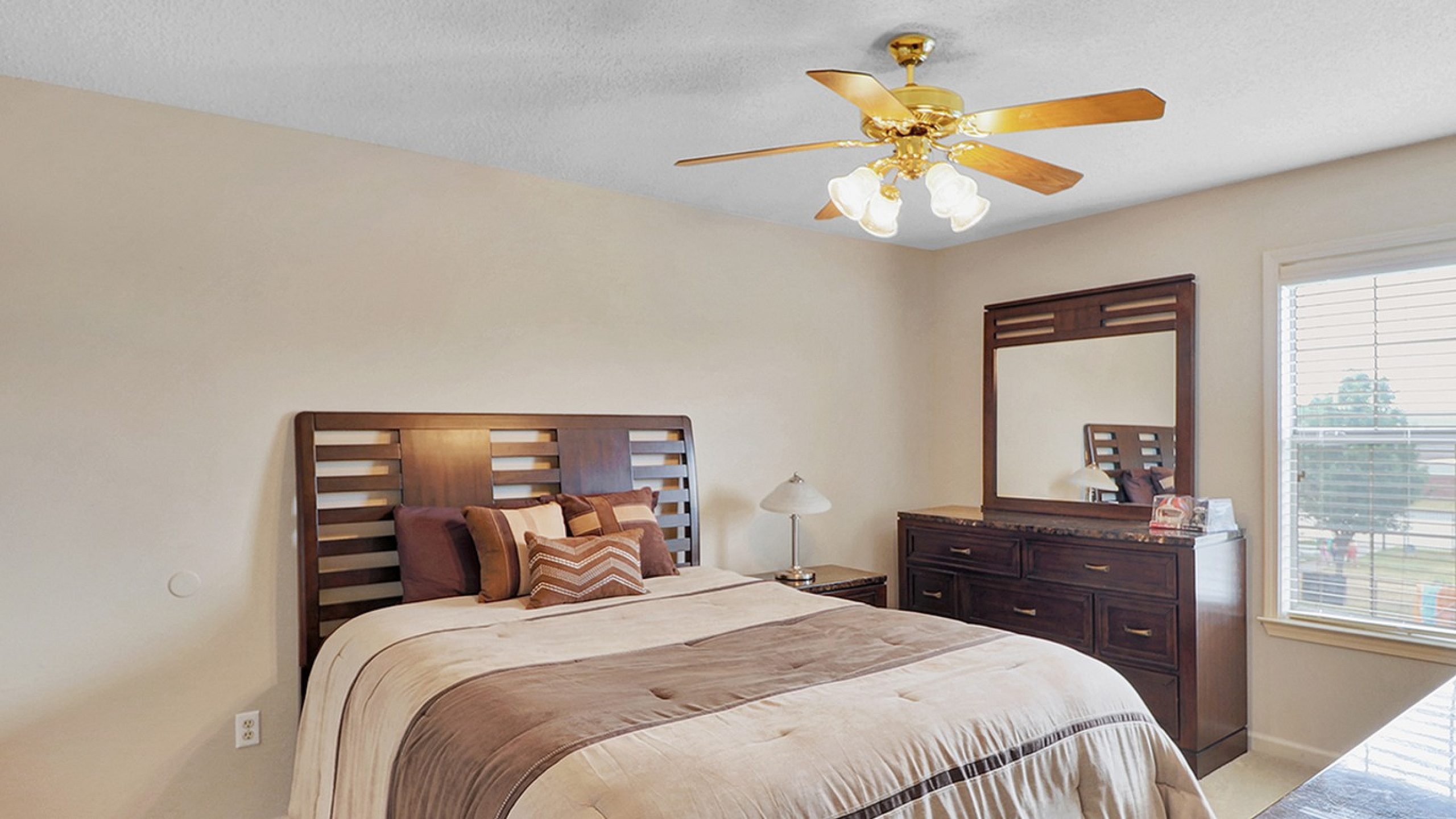 Cozy bedroom with elegant wooden furniture, featuring a large bed with plush bedding, complemented by a stylish ceiling fan and ample natural light.