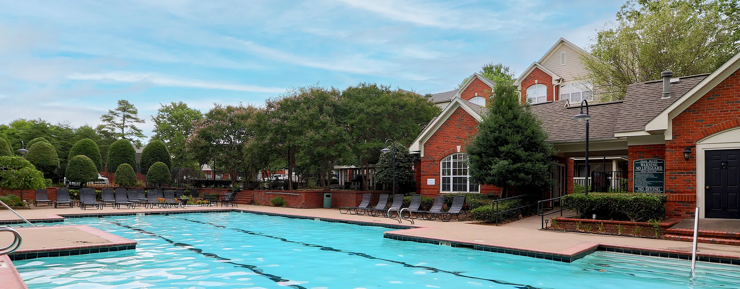 Inviting apartment complex pool with sun loungers, surrounded by lush greenery and elegant brick architecture under a bright blue sky.