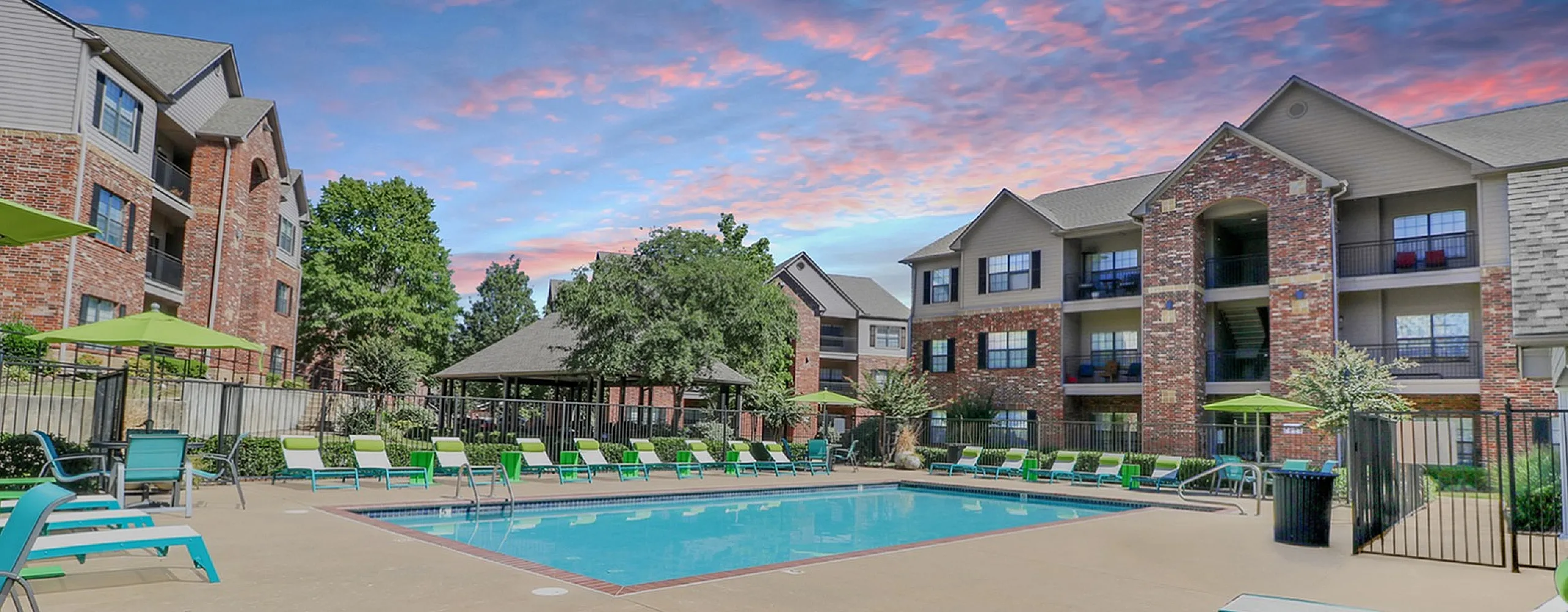 Charming apartment complex with a stunning pool area, surrounded by lush greenery and elegant architecture under a colorful sky.