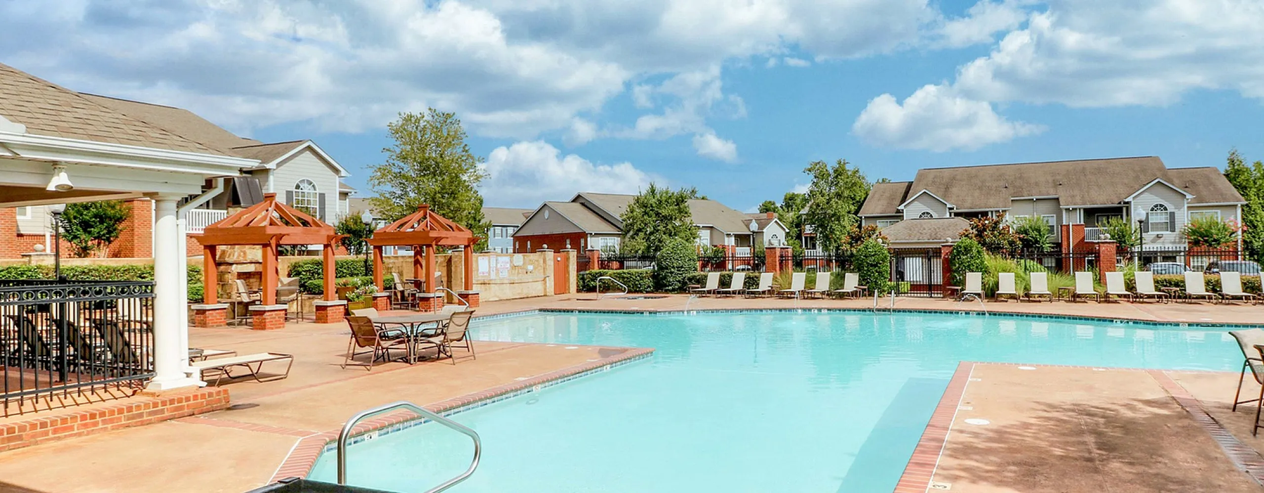 Luxurious apartment complex featuring a sparkling outdoor pool, stylish pergolas, and lush landscaping under a vibrant blue sky.