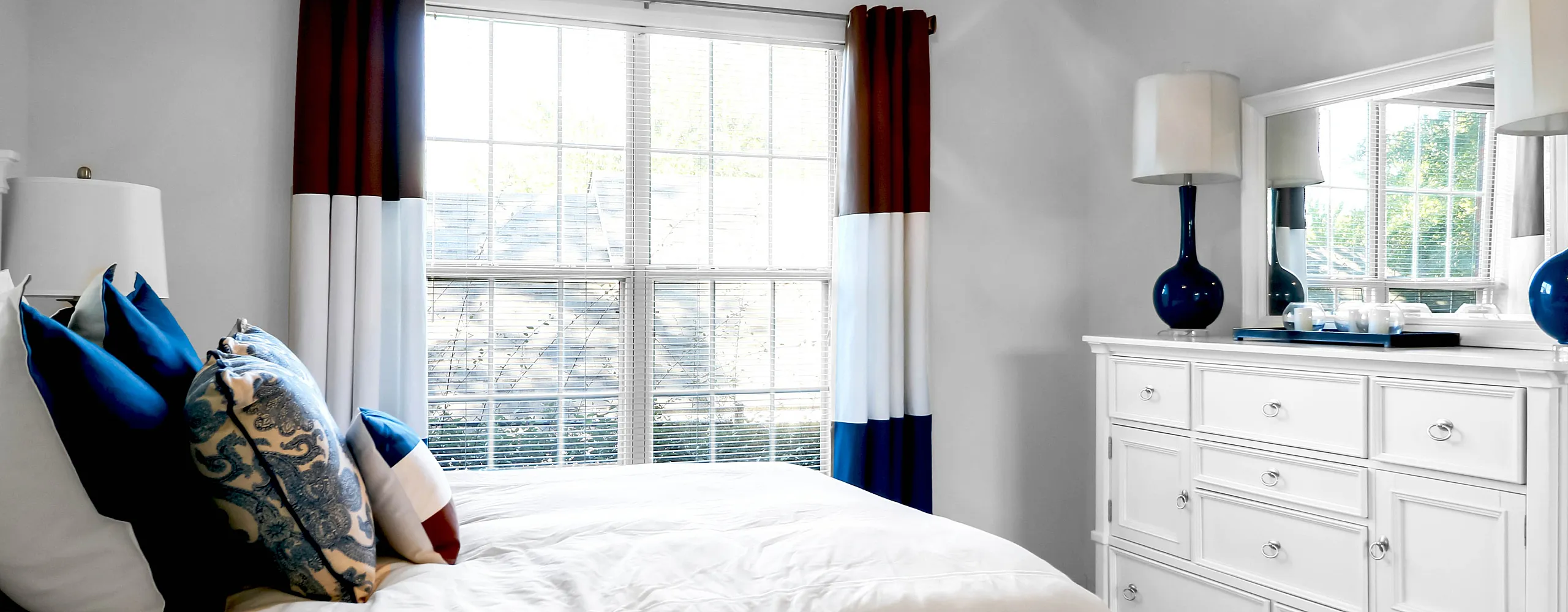 Bright, inviting bedroom with elegant white furniture, large windows with bold curtains, and cozy, plush pillows.