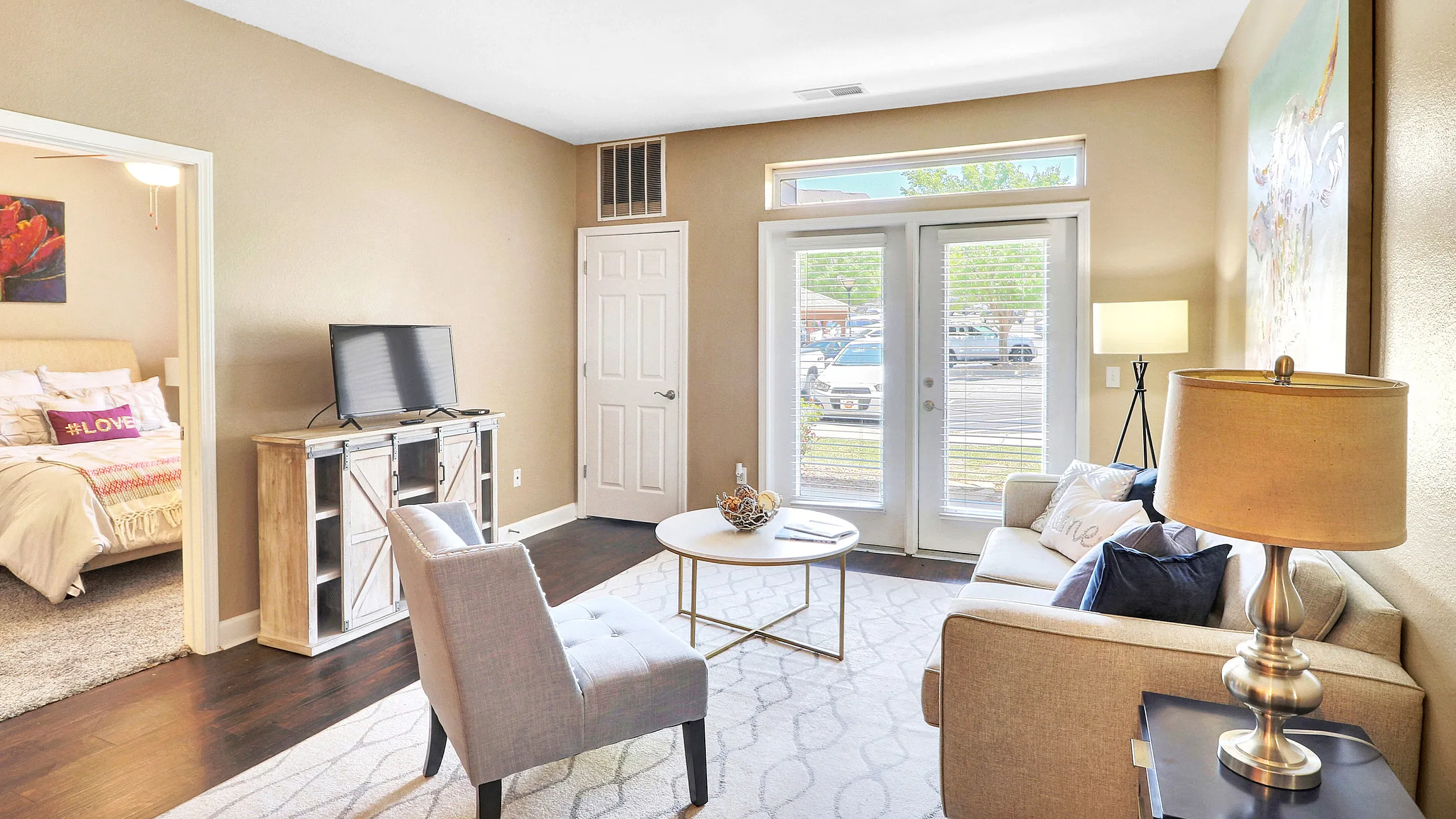 Inviting living room with elegant decor, featuring a cozy sofa, chic armchair, and stylish lamp, leading to a bedroom with a plush bed.