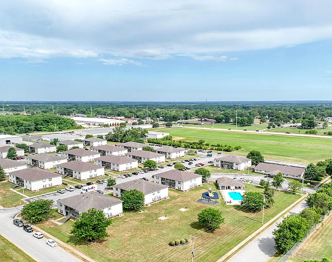 Aerial view of a beautifully landscaped apartment complex with a sparkling pool, abundant green spaces, and convenient parking, set against a serene backdrop of lush greenery and open skies.