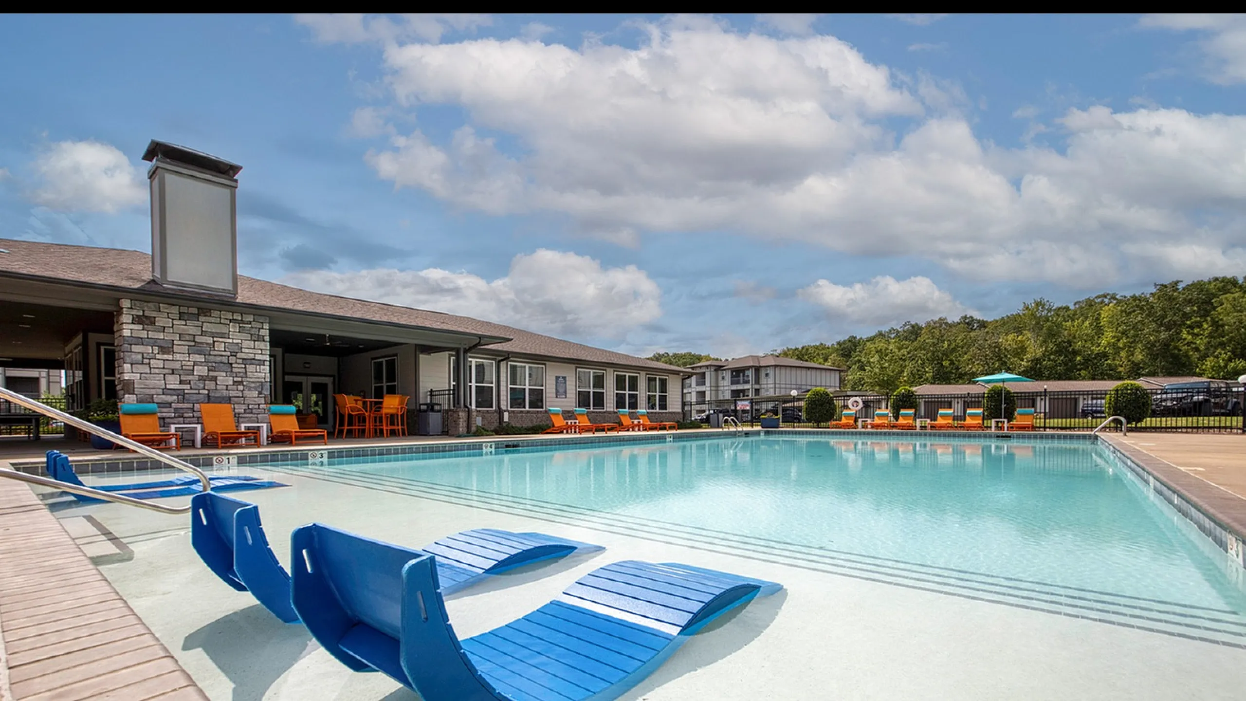 Luxurious outdoor pool area featuring modern blue lounge chairs and vibrant orange seating, set against a stylish clubhouse exterior surrounded by lush greenery.